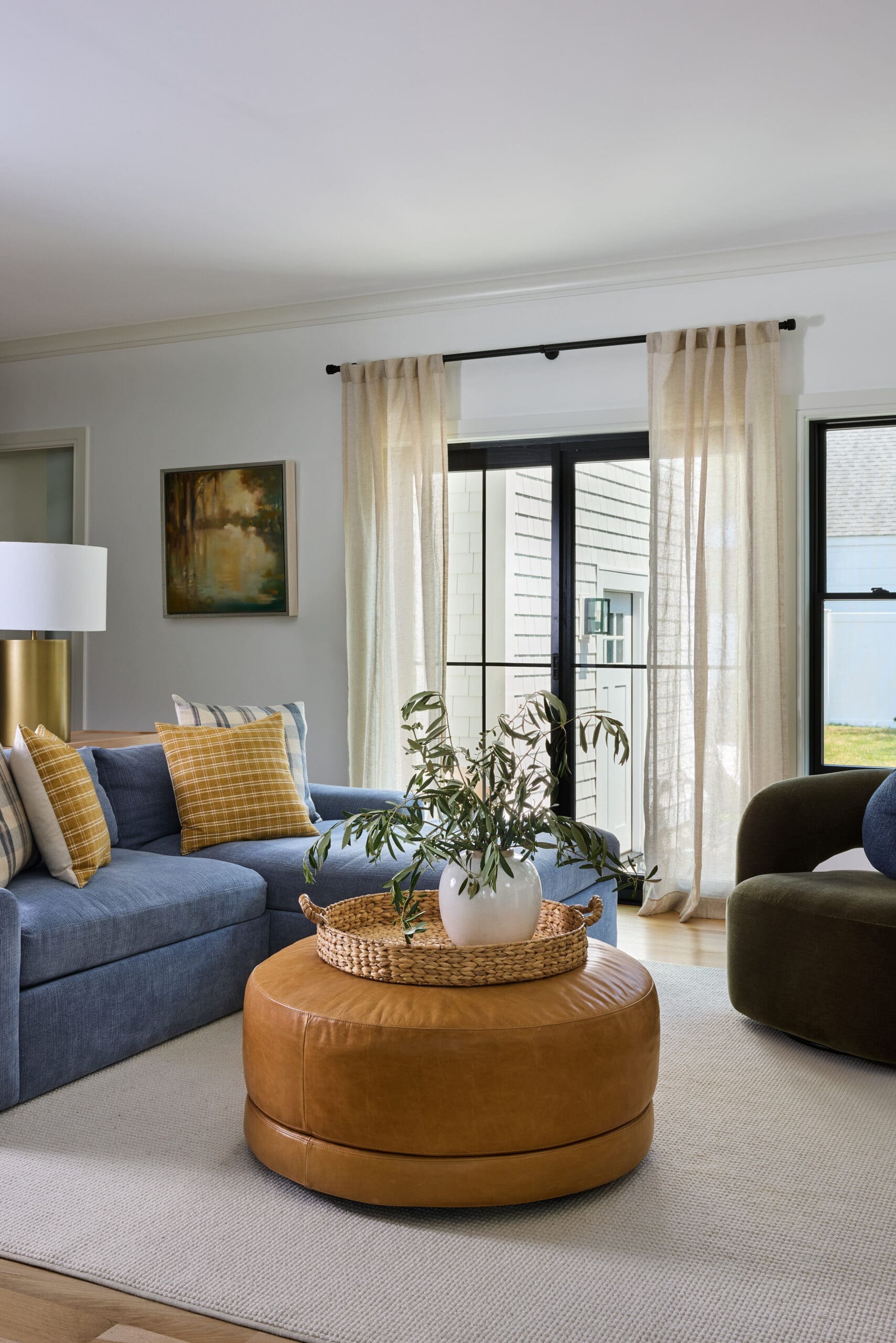 Cozy living room with a blue sectional sofa accented by mustard plaid cushions, a round tan leather ottoman with a woven tray and plant, beige sheer curtains, and large windows bringing in natural light
