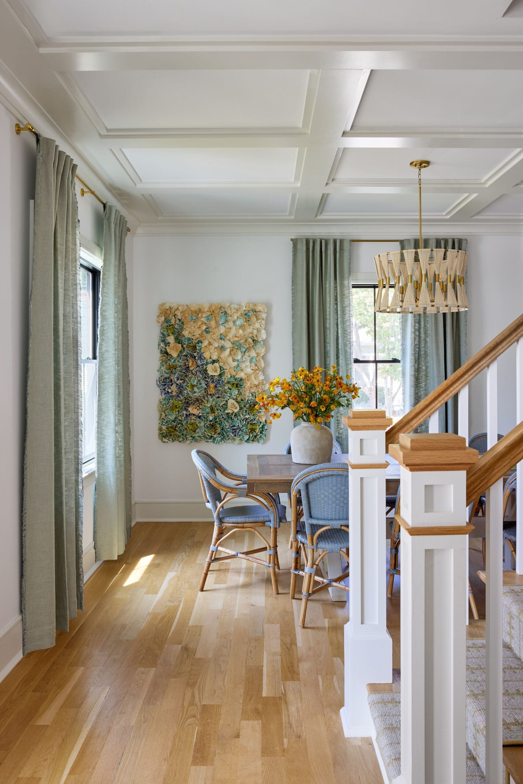 Bright dining area with a wooden table, blue chairs, and a woven chandelier; light pours from windows with green curtains.