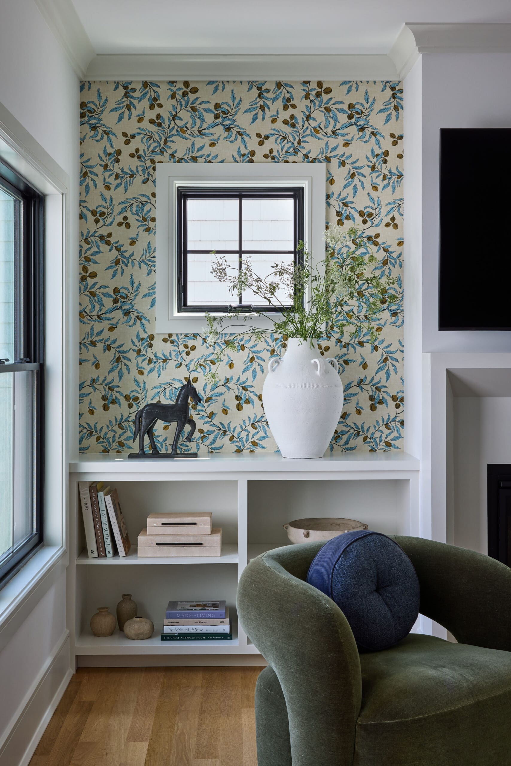 Interior nook with blue botanical wallpaper, a square window, a white vase with greenery, and a small dark horse statue on a shelf.