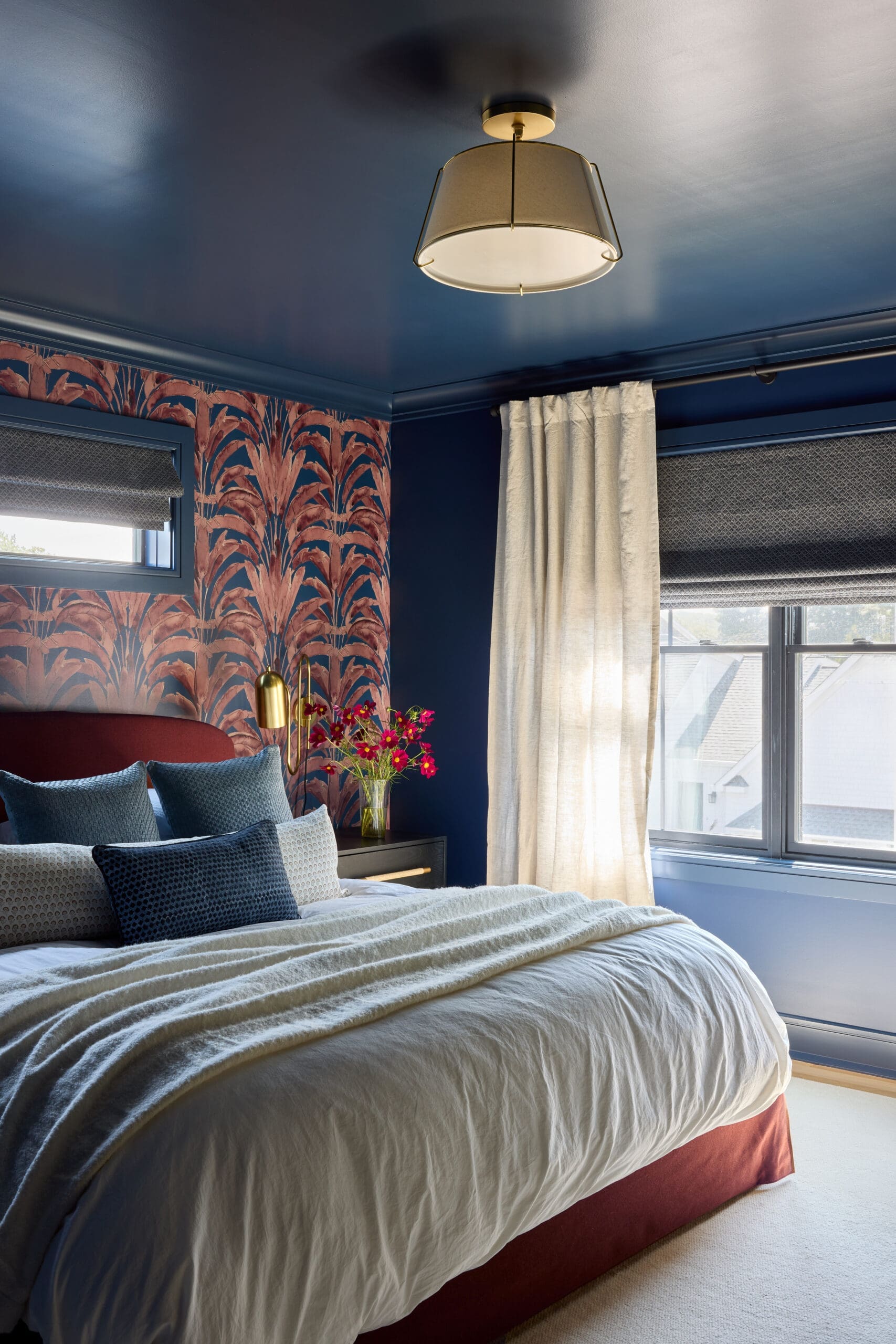 Bedroom with navy walls, pink tropical wallpaper, and a white duvet on a red upholstered bed. Window with blinds to the right of sheer curtains.