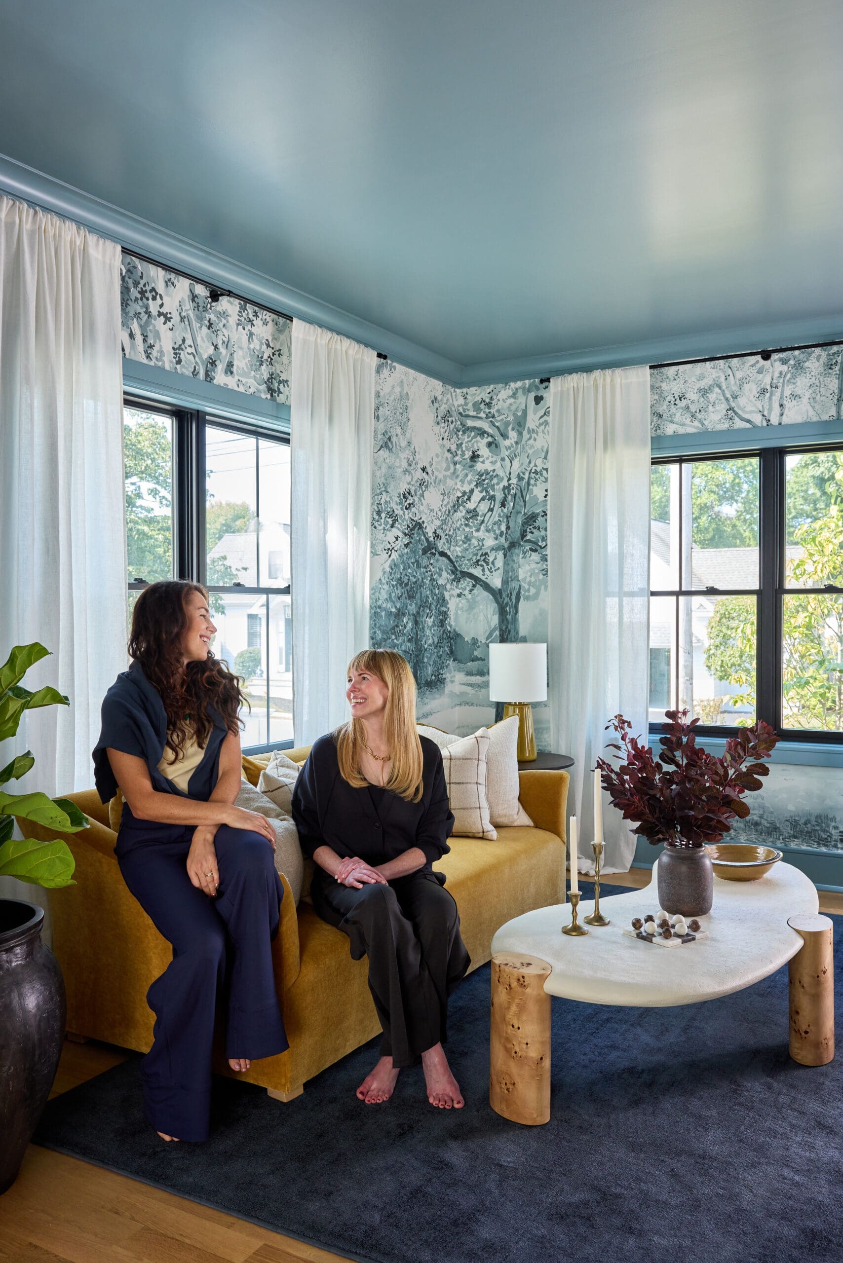 Two women sit and chat on a mustard sofa in a bright living room with blue walls, sheer curtains, and botanical wallpaper.
