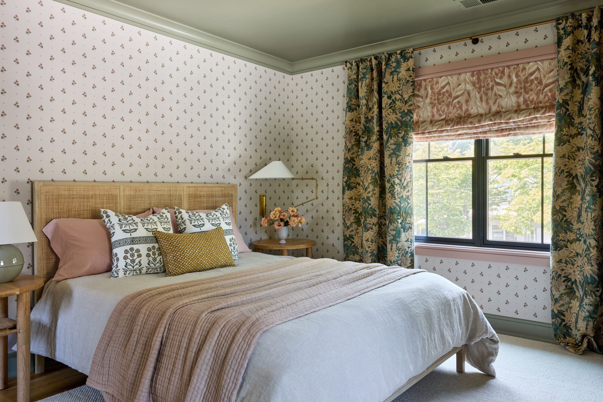 Cozy bedroom with a woven headboard, beige quilt, and pink pillows, floral wallpaper, and a window with patterned curtains.