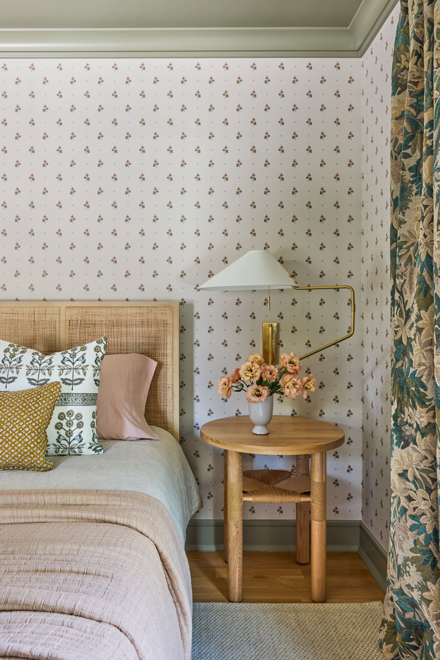 Cozy bedroom corner with a wicker headboard, pastel bedding, and decorative pillows, beside a round wooden bedside table with a vase of pink flowers and a brass lamp against floral wallpaper.