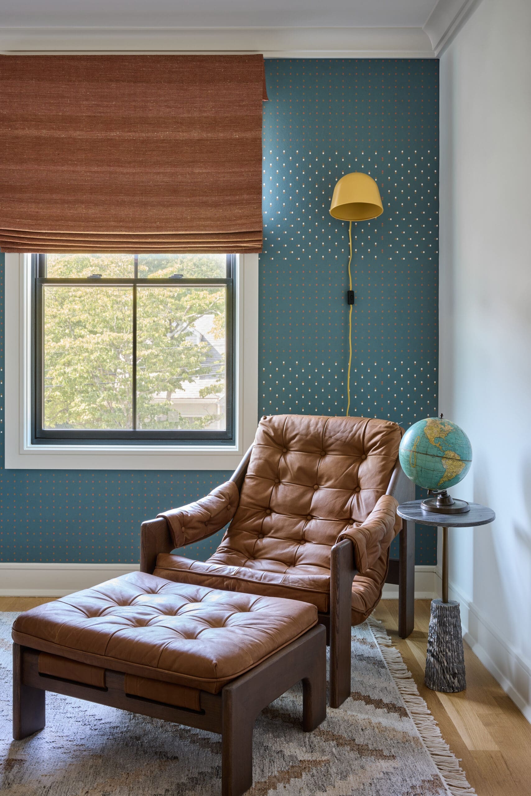 Cozy reading nook with a brown leather tufted chair and matching ottoman, blue dotted wallpaper, and a yellow wall lamp beside a window with brown blinds and a globe on a side table.