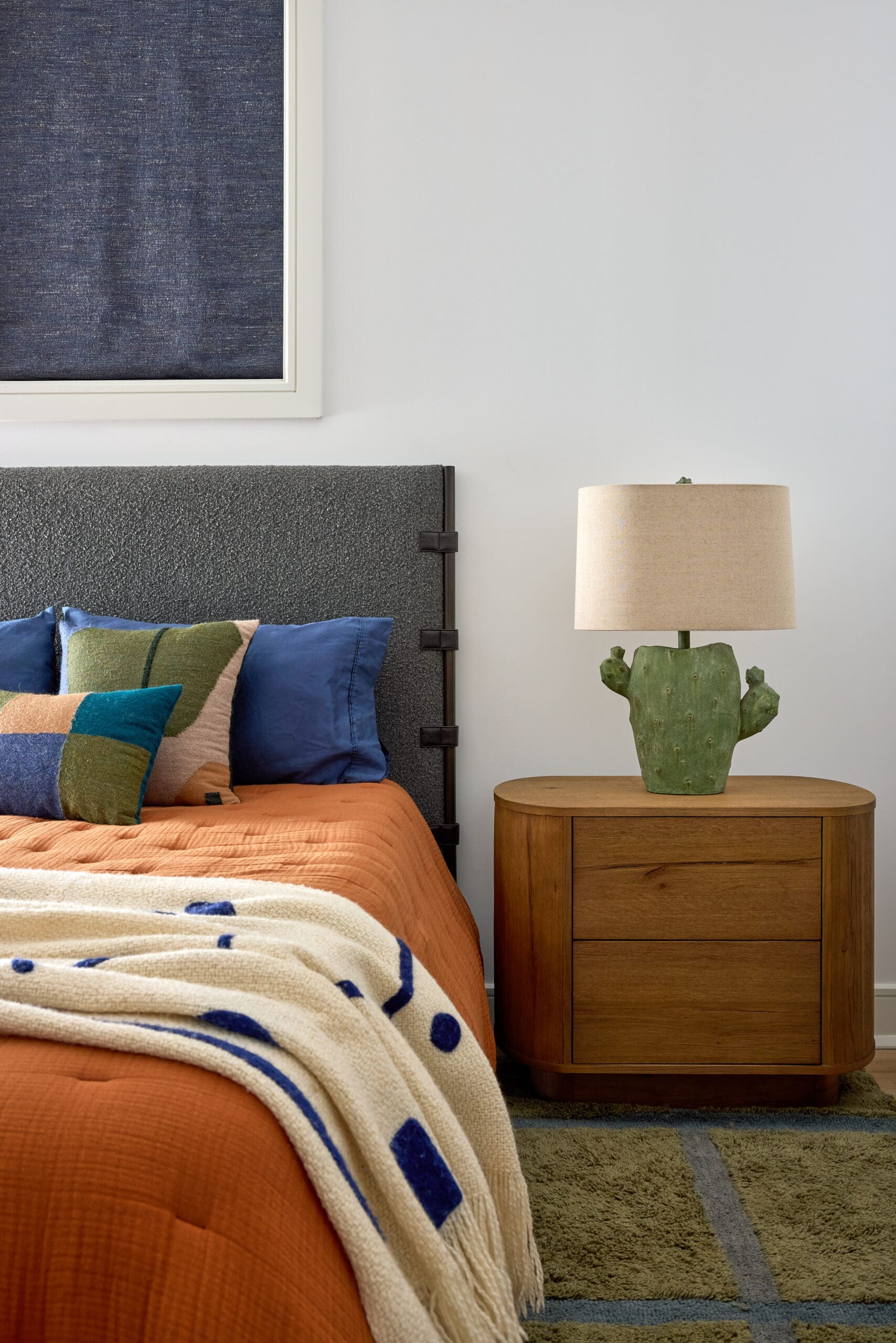 Bedroom scene with an orange quilt, colorful pillows, and a textured dark headboard beside a wooden nightstand with a beige lamp and a cactus plant.