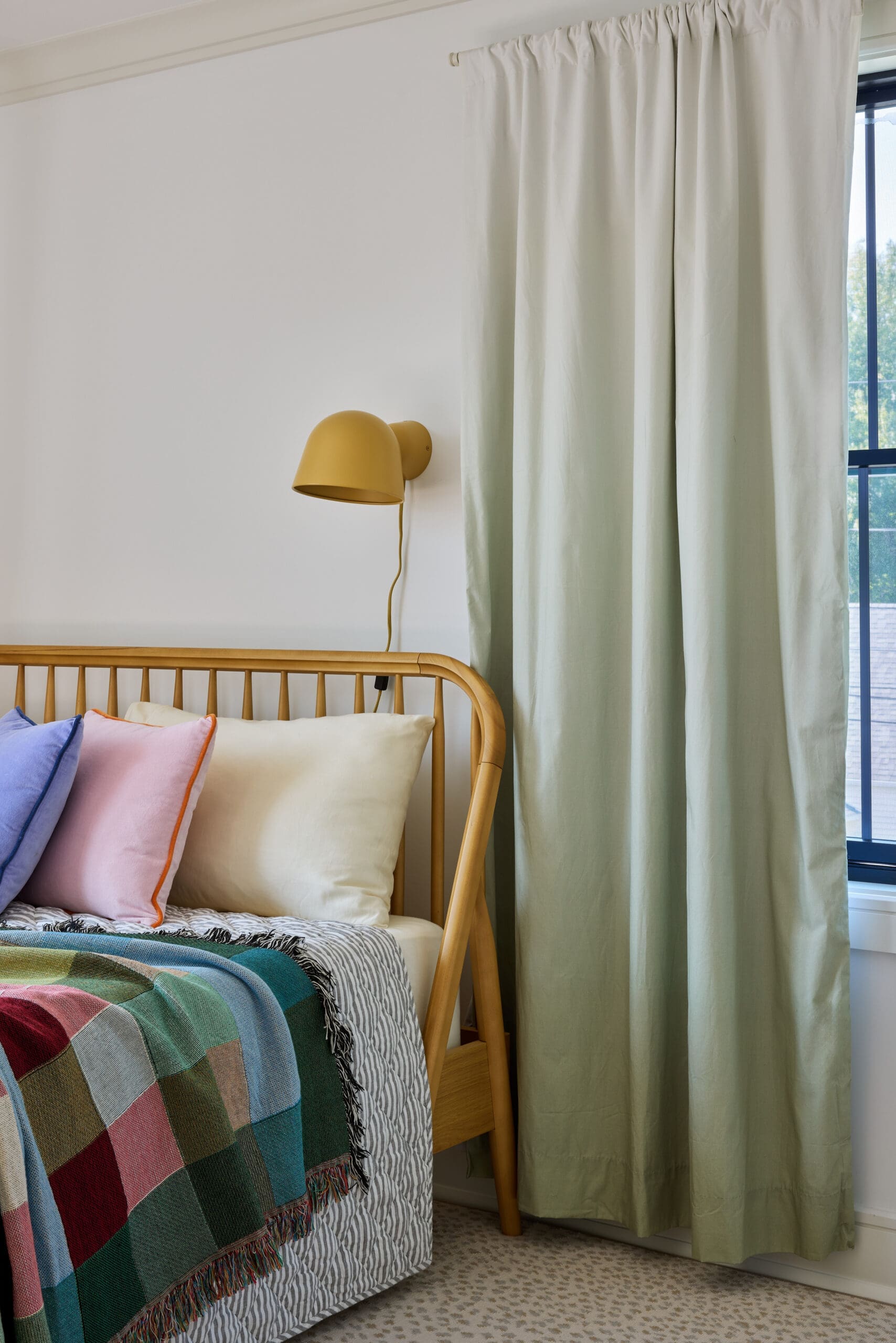 Wooden bed with pastel pillows and a multicolored plaid blanket, beside a mustard wall lamp and pale green curtains in a bright bedroom corner.