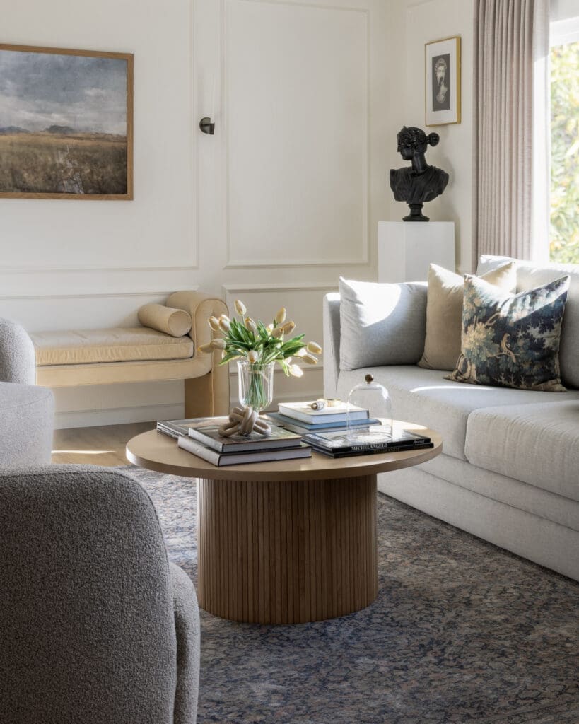 Living room vignette with a round ribbed-wood coffee table, vase of tulips, beige seating, and a black bust sculpture on a pedestal by the window.