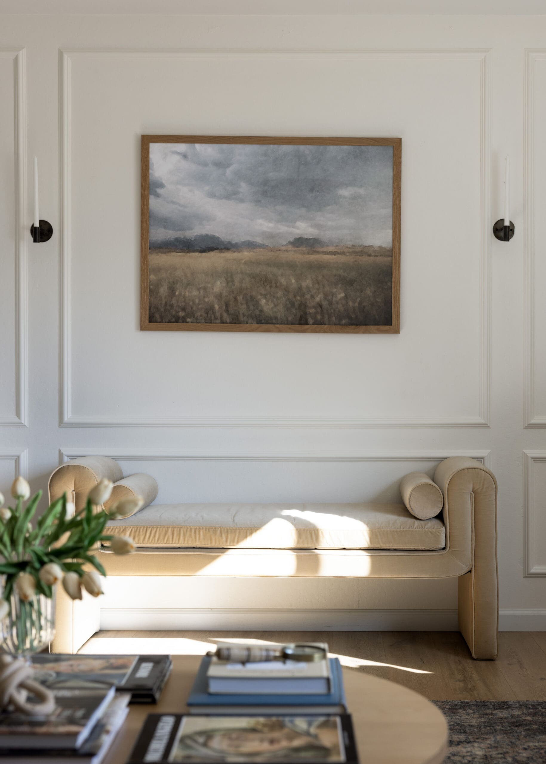 Framed landscape painting on a white paneled wall above a beige sofa with cushions in a cozy living room.