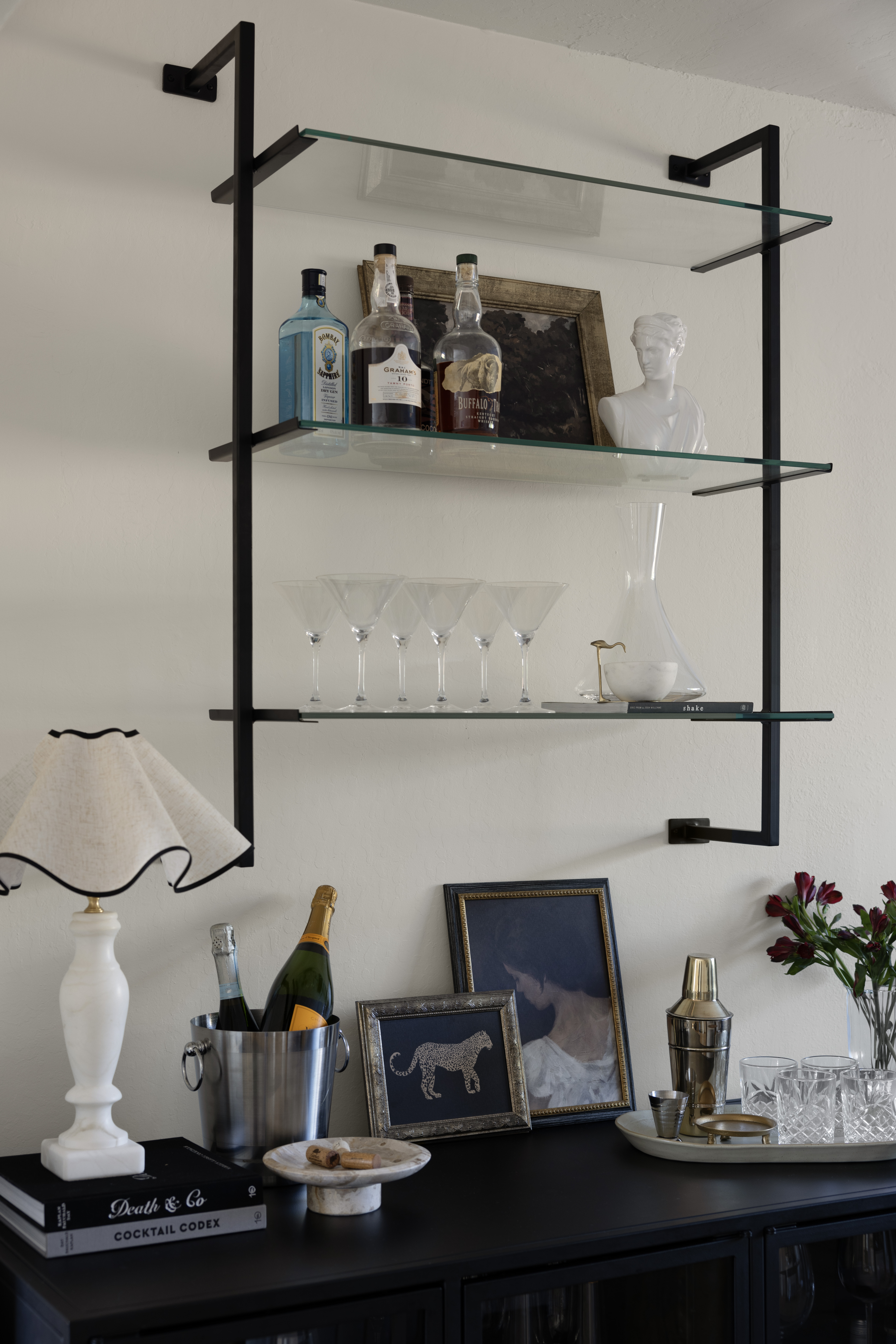 Wall-mounted black metal and glass shelf holding liquor bottles, a white bust statue, and framed art behind. (Bar display)
