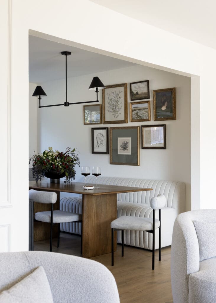 Dining nook with a wooden table, upholstered bench, and a black multi-arm chandelier; wall gallery of framed art pieces above the seating area.