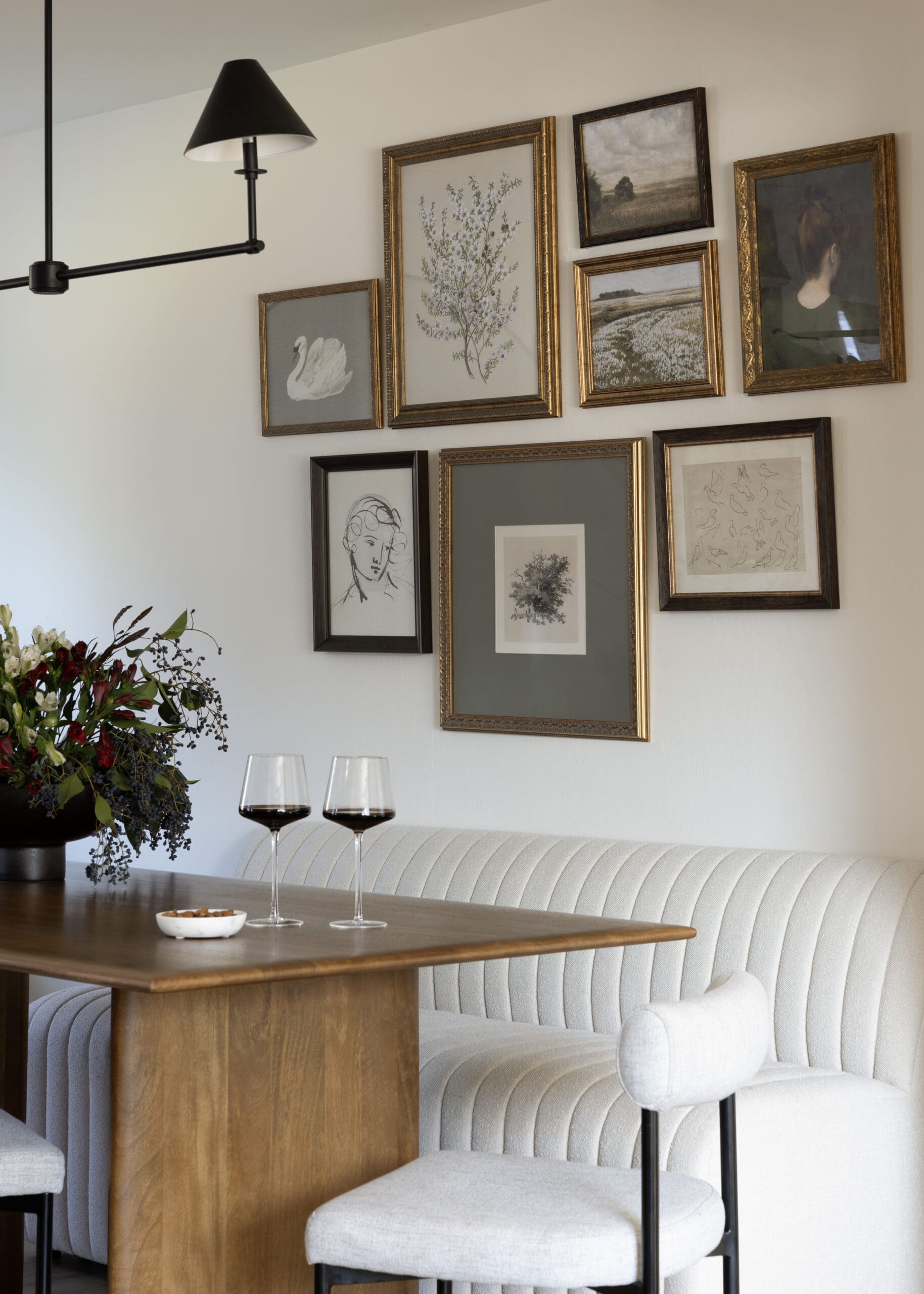 Dining area with a wall of framed artwork above a tufted white bench and a wooden table; two wine glasses sit on the table, and a bouquet is on the left.