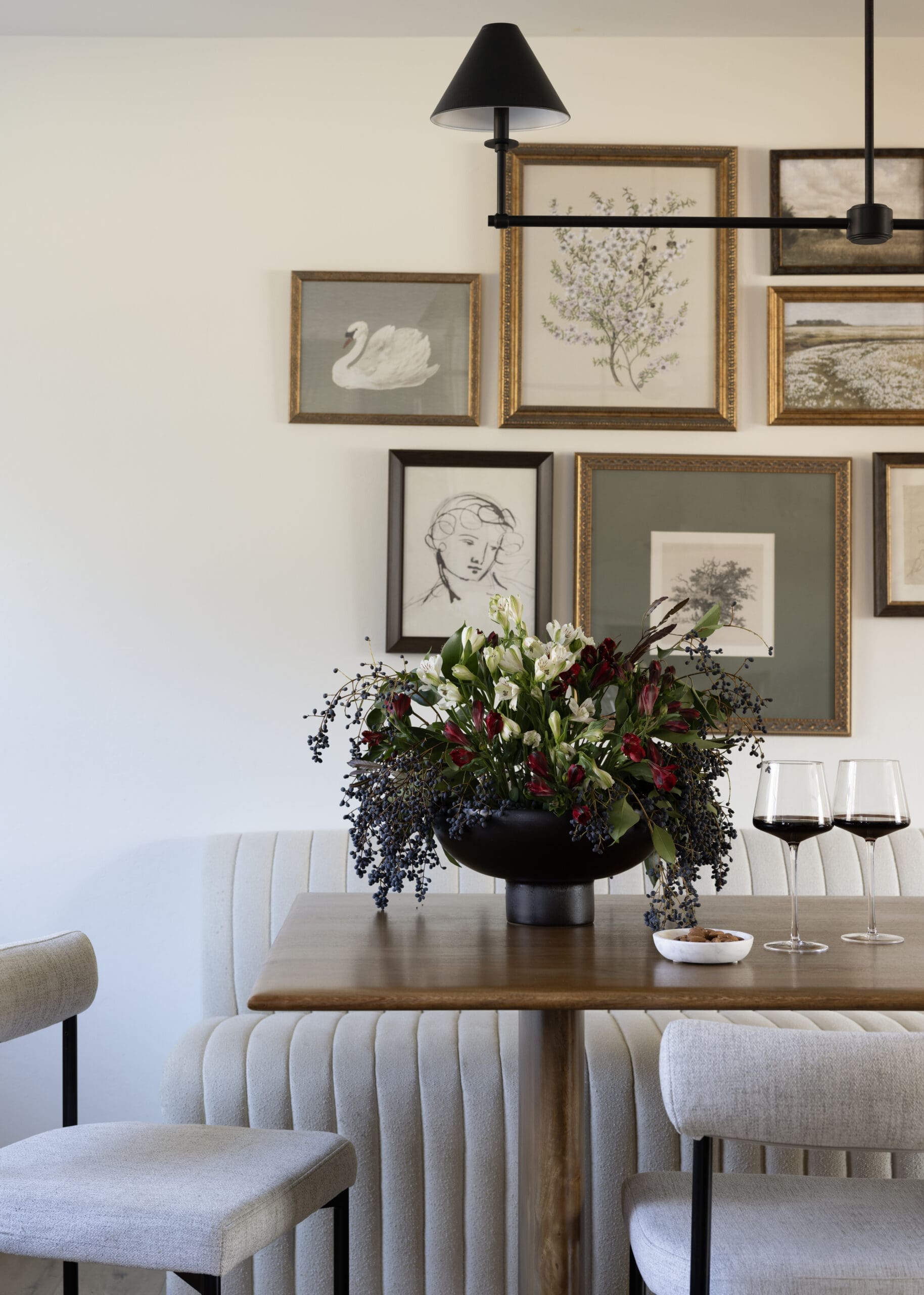 A dining area with a dark vase of white and red flowers on a wooden table, framed art on the wall, and two wine glasses nearby.