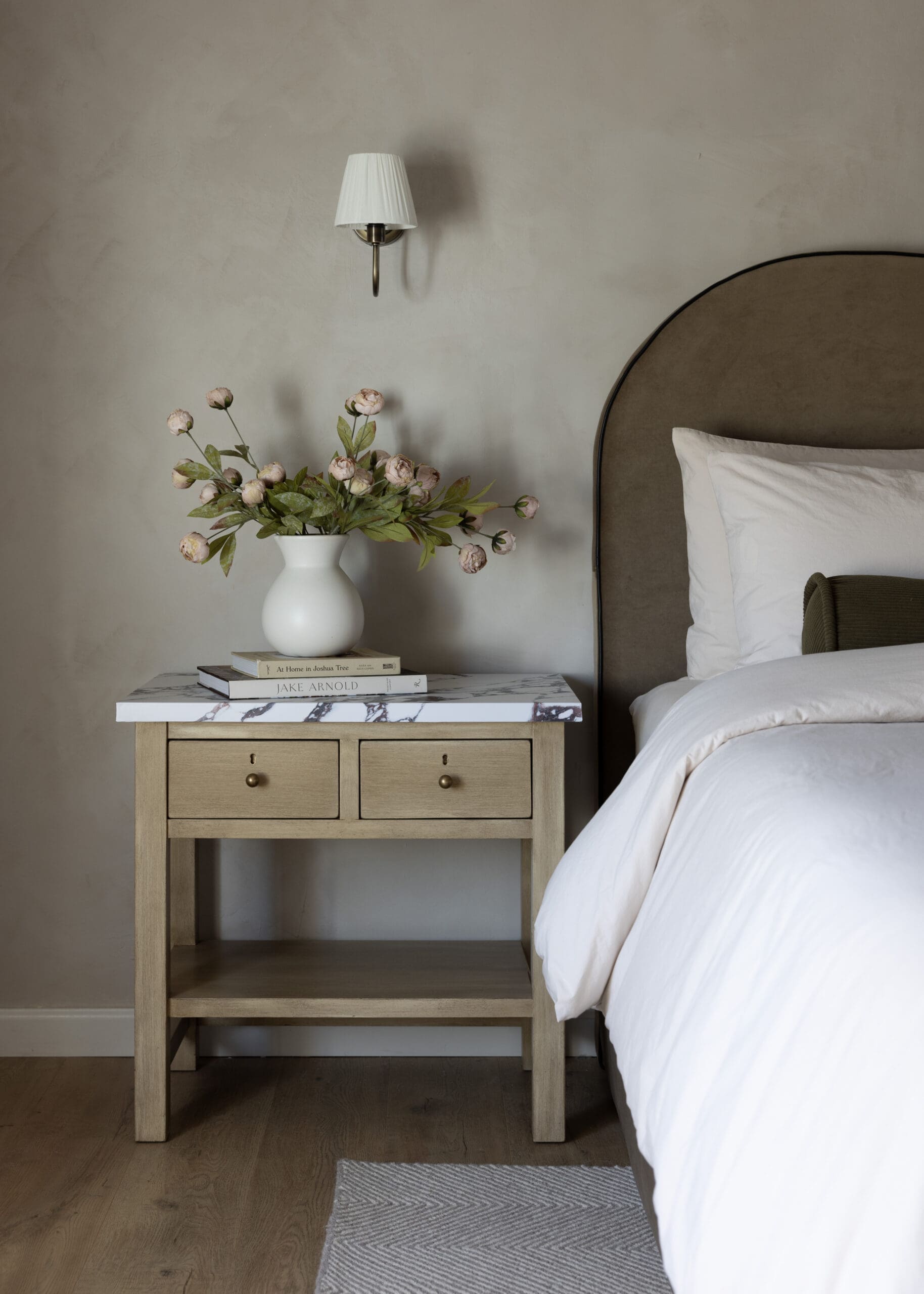 Beige upholstered headboard with white bedding, and a wooden nightstand topped with a marble surface, a white vase of pink peonies, and books.