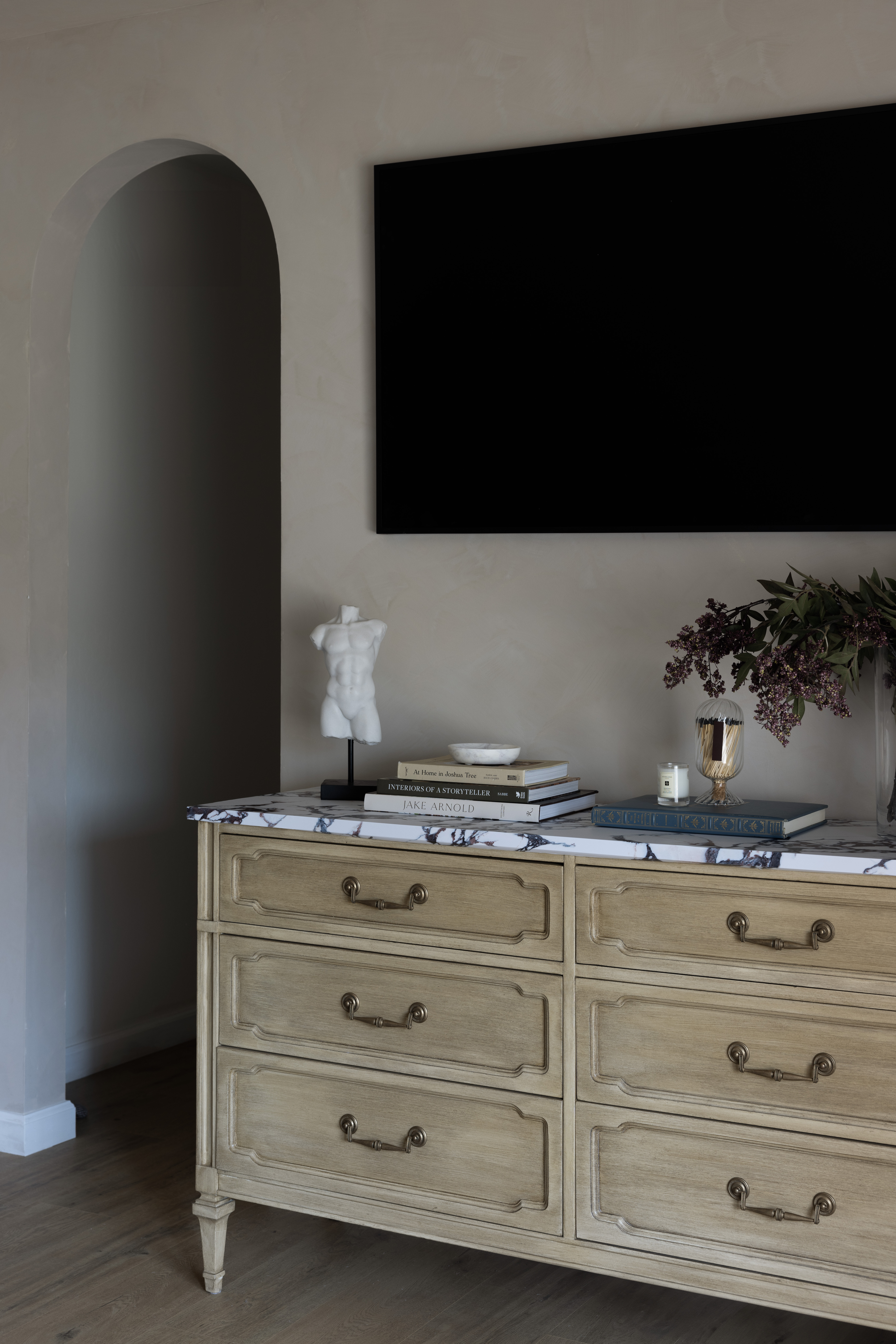 Beige dresser with marble top, white torso sculpture on a stand, stacked books, a vase of purple flowers, and a large black TV on the wall above.