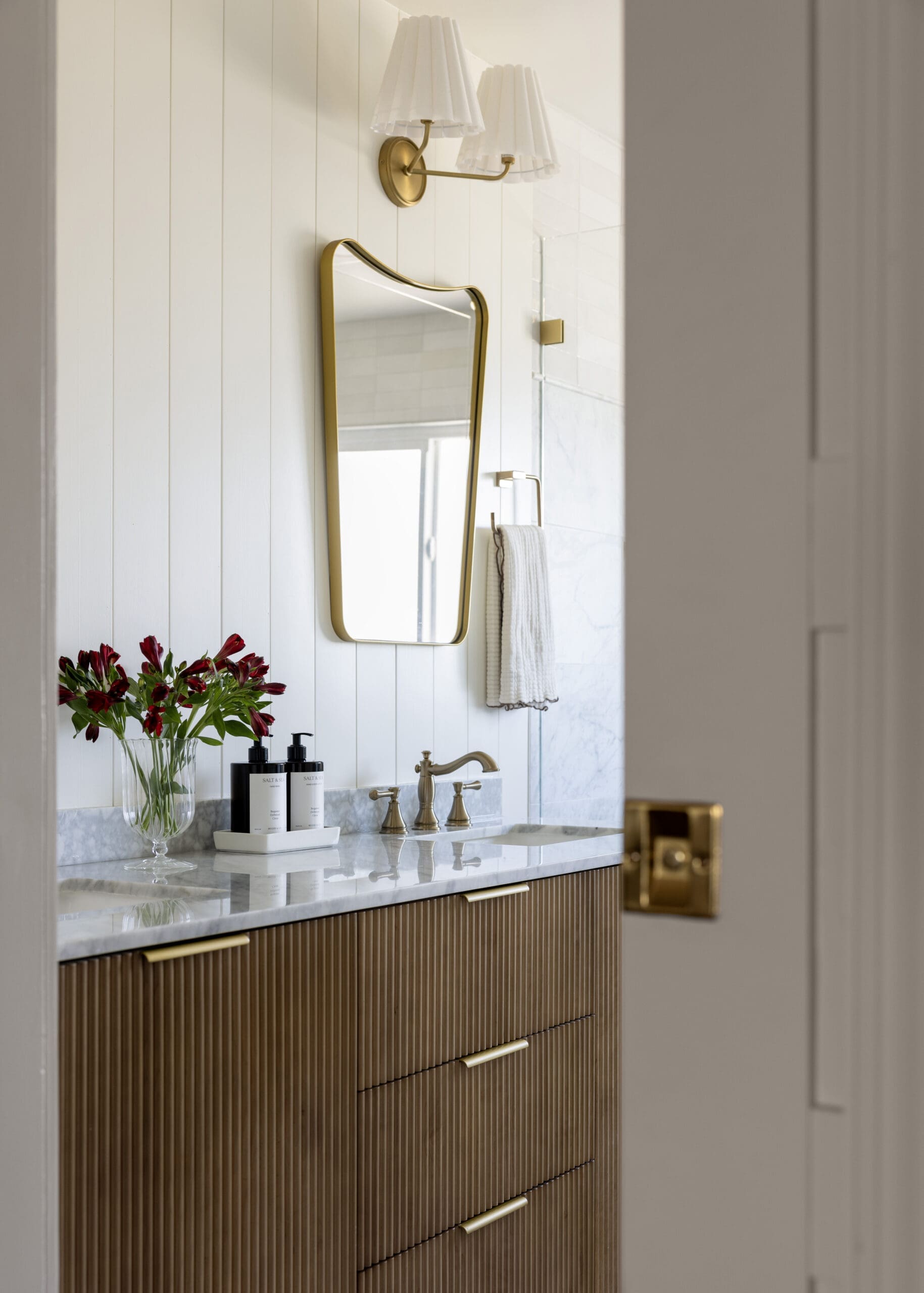 Bathroom vanity with gold-brass faucet, curved framed mirror, white beadboard walls, and a vase of red flowers on a marble countertop.