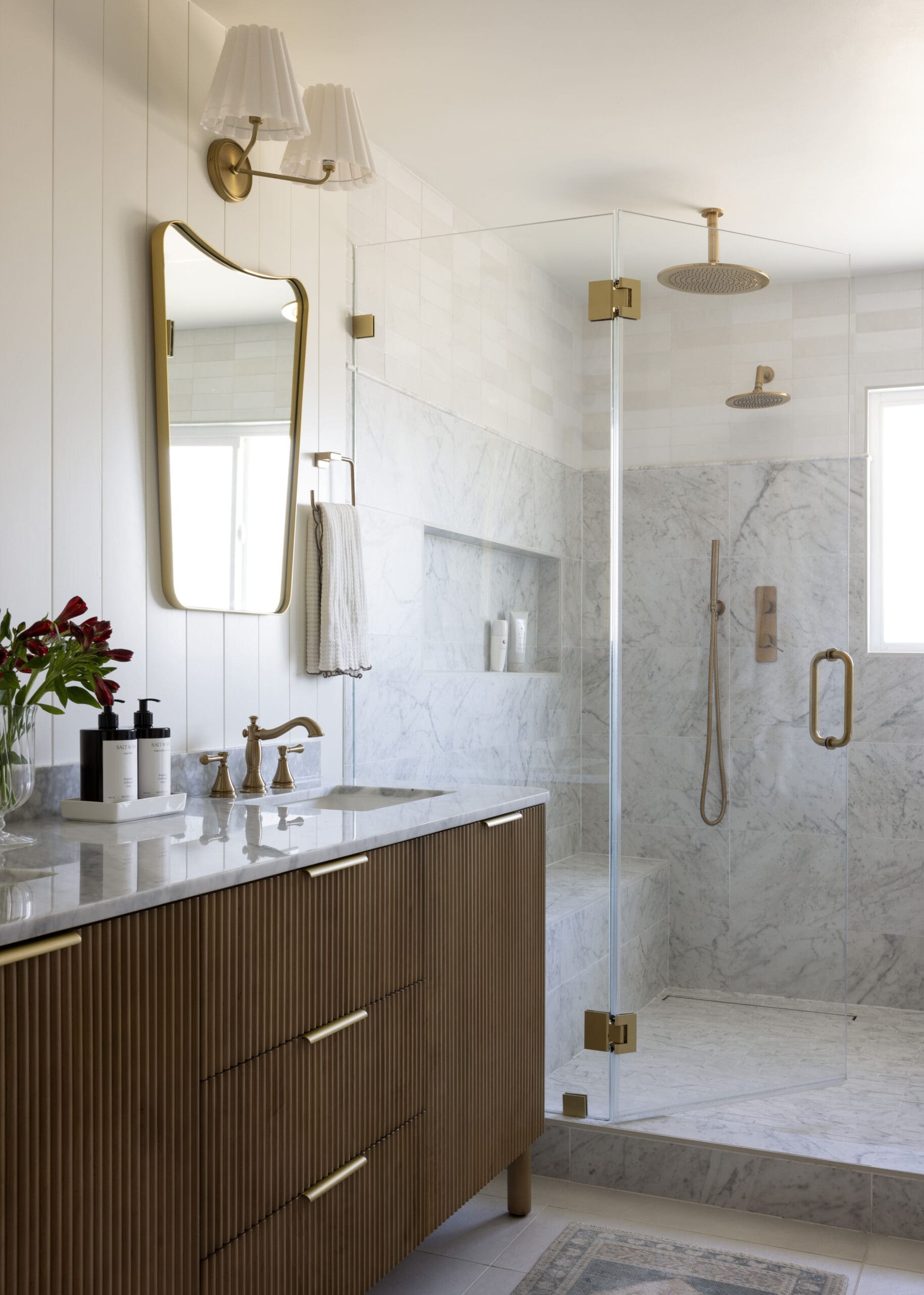 Luxurious bathroom with a marble vanity, gold fixtures, and a glass-walled shower featuring a rainfall head and handheld sprayer, plus a gold-framed mirror and vase of red flowers on the counter.