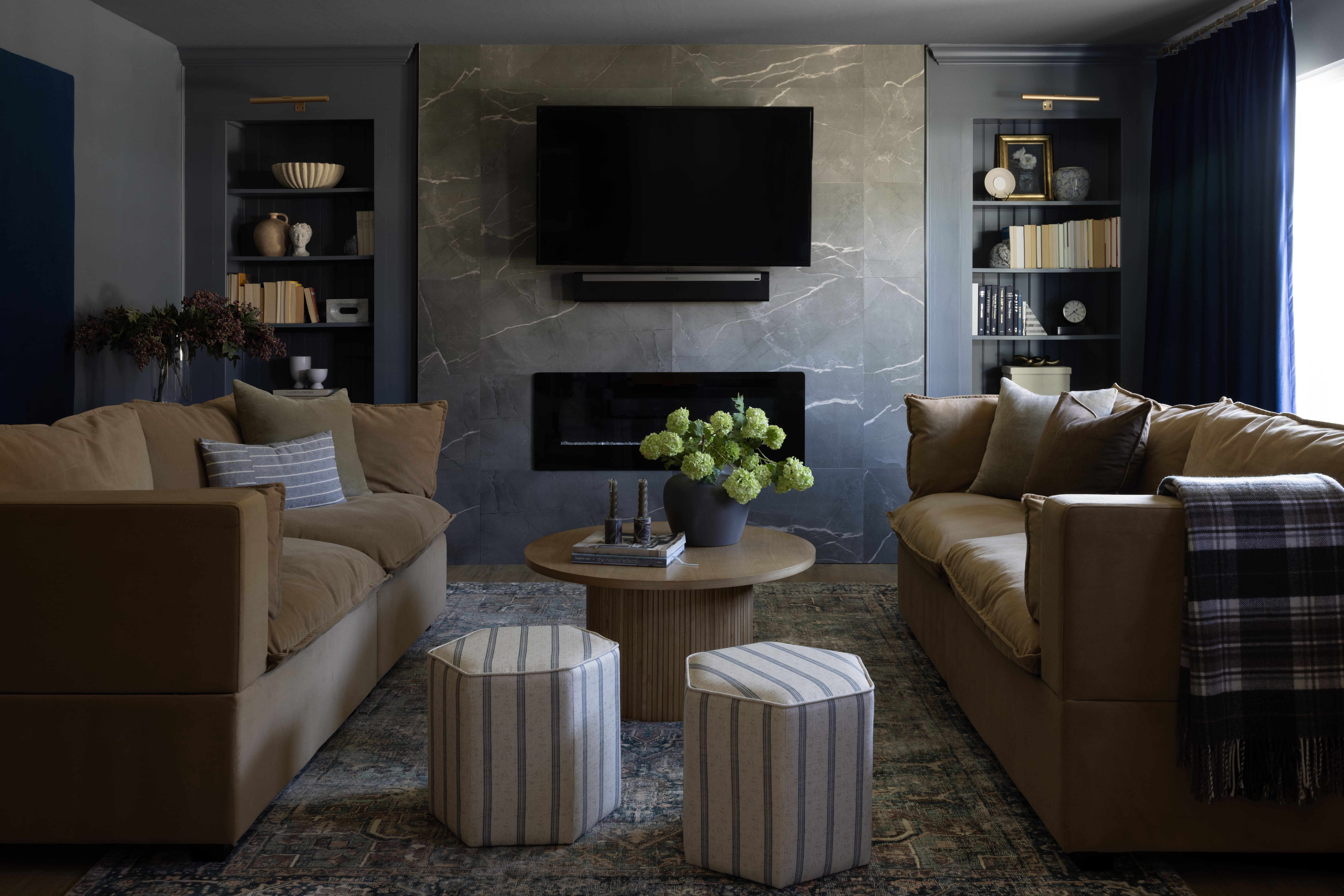Two tan sectional sofas face a central marble-feature wall with a mounted flat-screen TV and built-in blue bookshelves on either side.