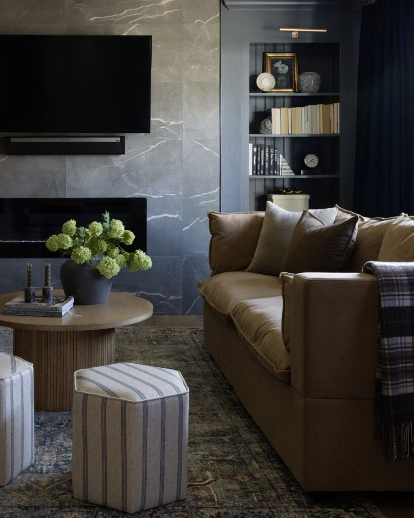 Living room scene with a round wooden coffee table, a vase of green hydrangeas, and a tan sofa in front of a marble wall and built‑in bookshelf.