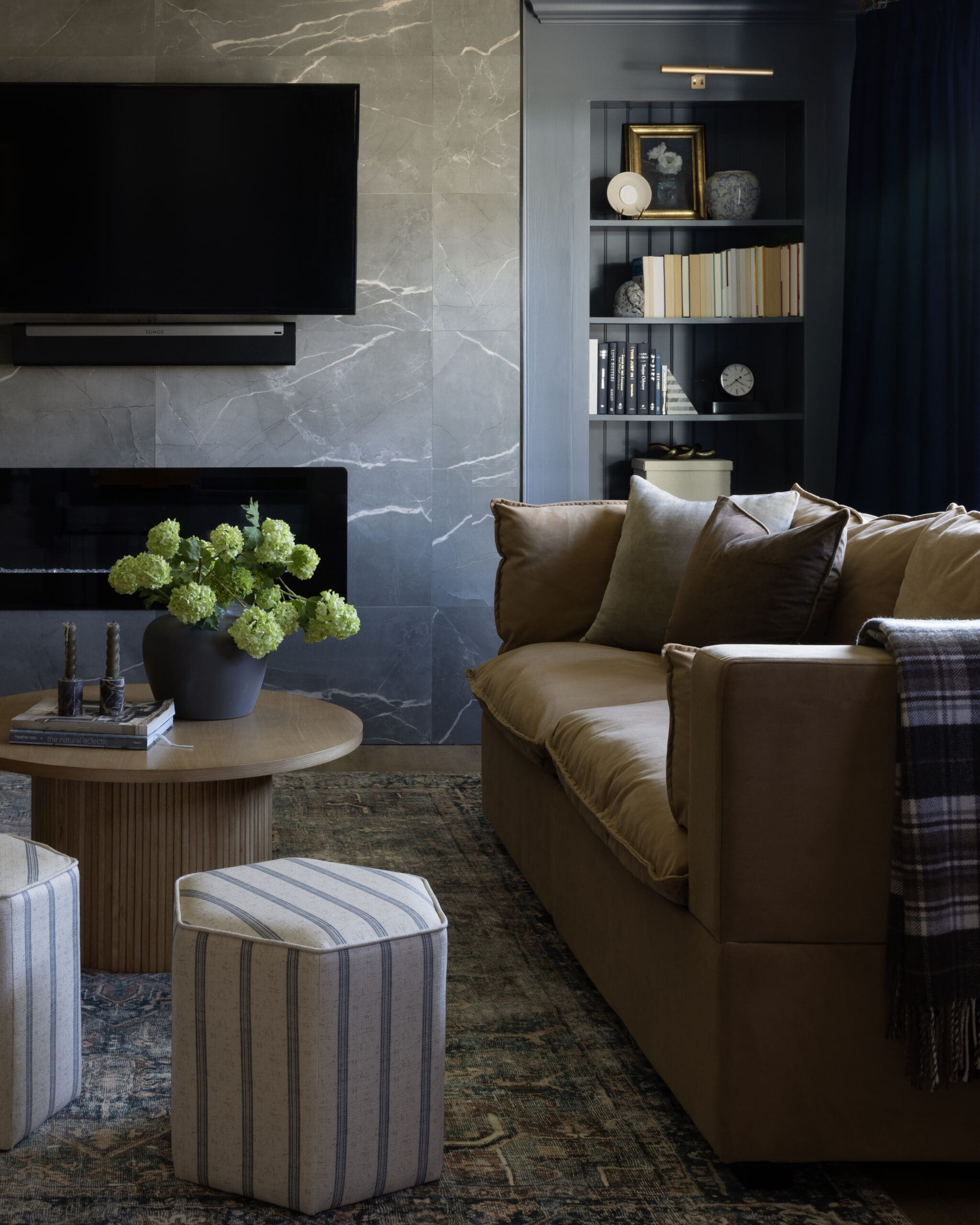 Living room scene with a round wooden coffee table, a vase of green hydrangeas, and a tan sofa in front of a marble wall and built‑in bookshelf.