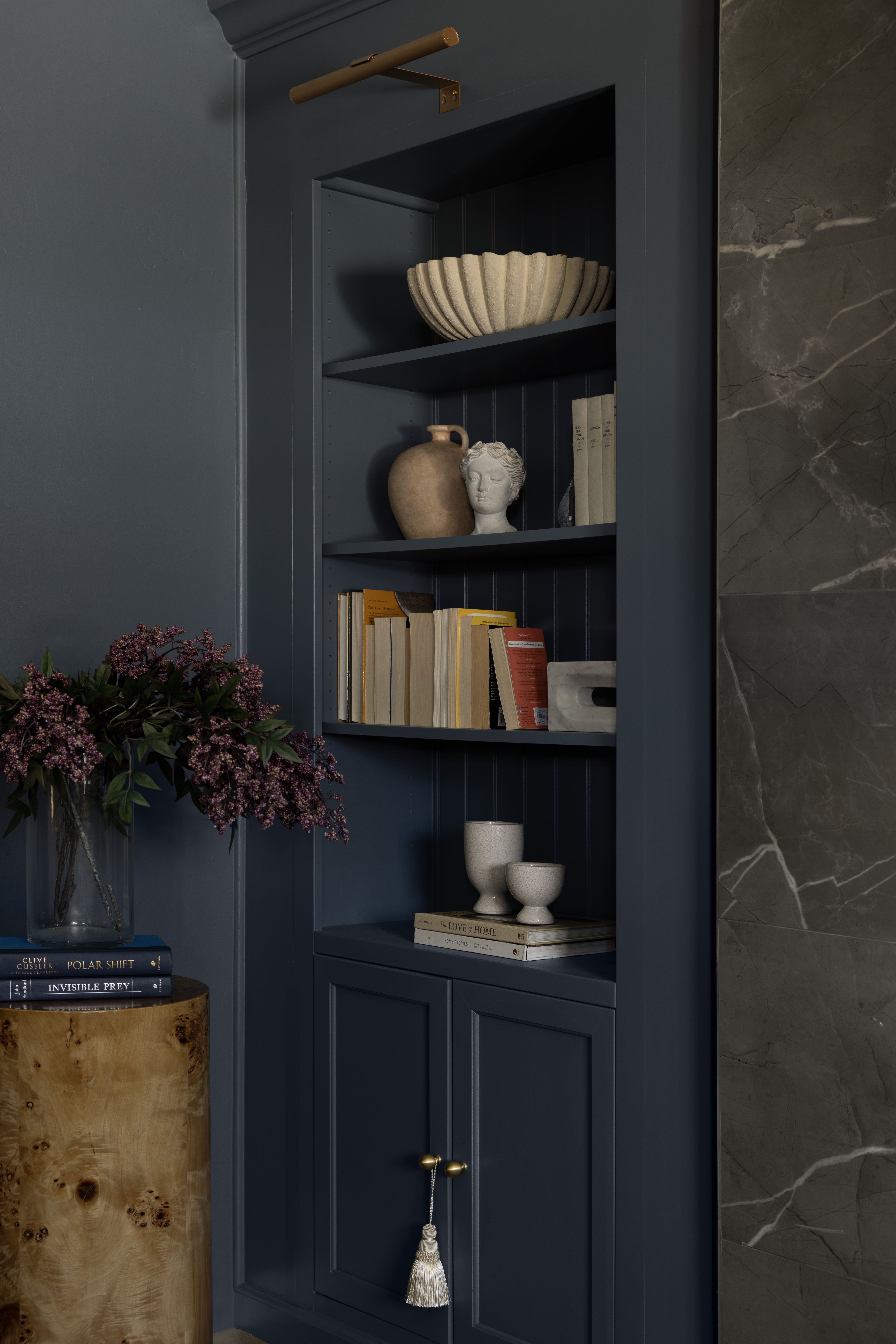Built-in dark blue cabinet with open shelves holding a ribbed beige bowl, a stone bust, books, a clay jug, and ceramic cups; a tassel hangs from the lower door handle beside a marble wall edge.