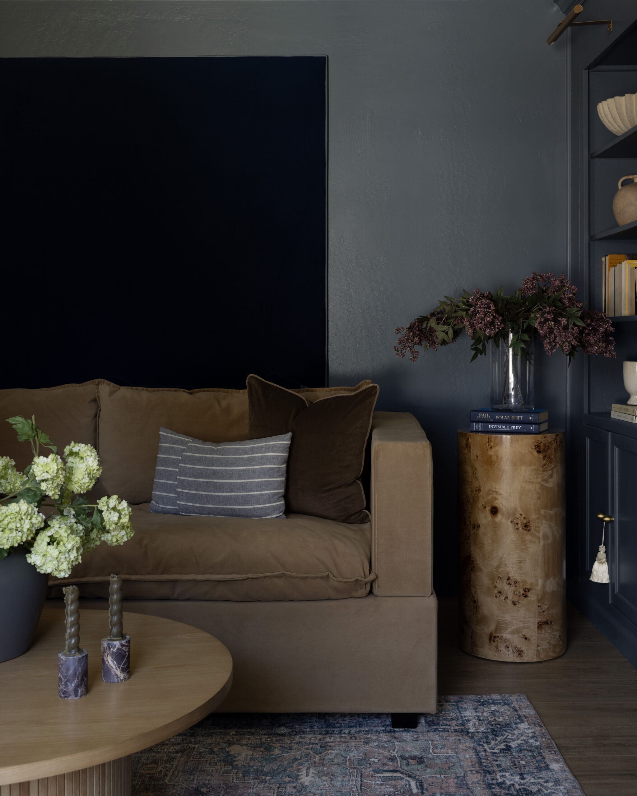 Beige sofa with brown cushions beside a navy wall, round wooden coffee table displaying a floral arrangement and three decorative candles at the edge of the room.