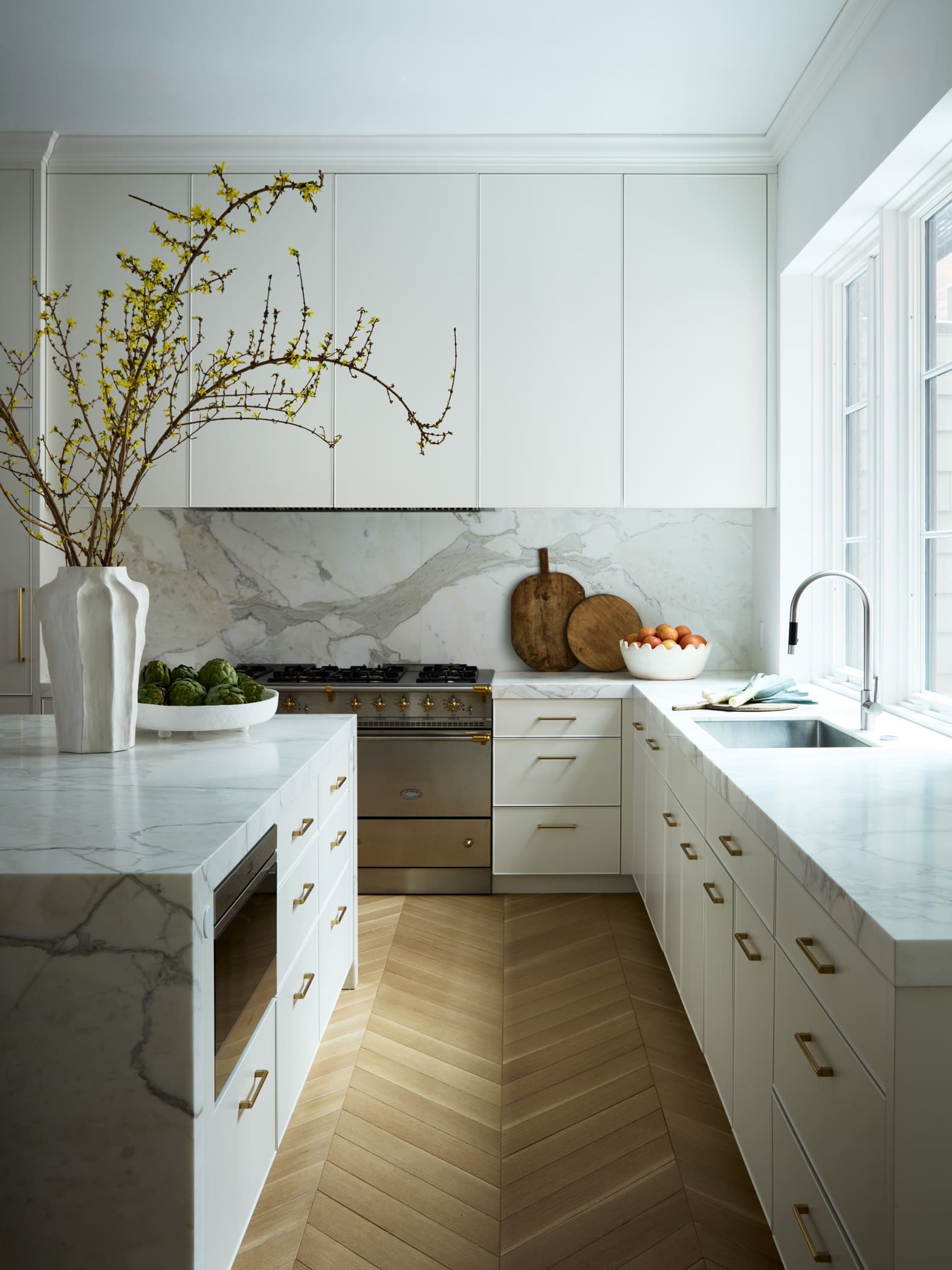 Bright white kitchen with marble countertops and a vase of flowering branches on the island, near a windowed sink area.