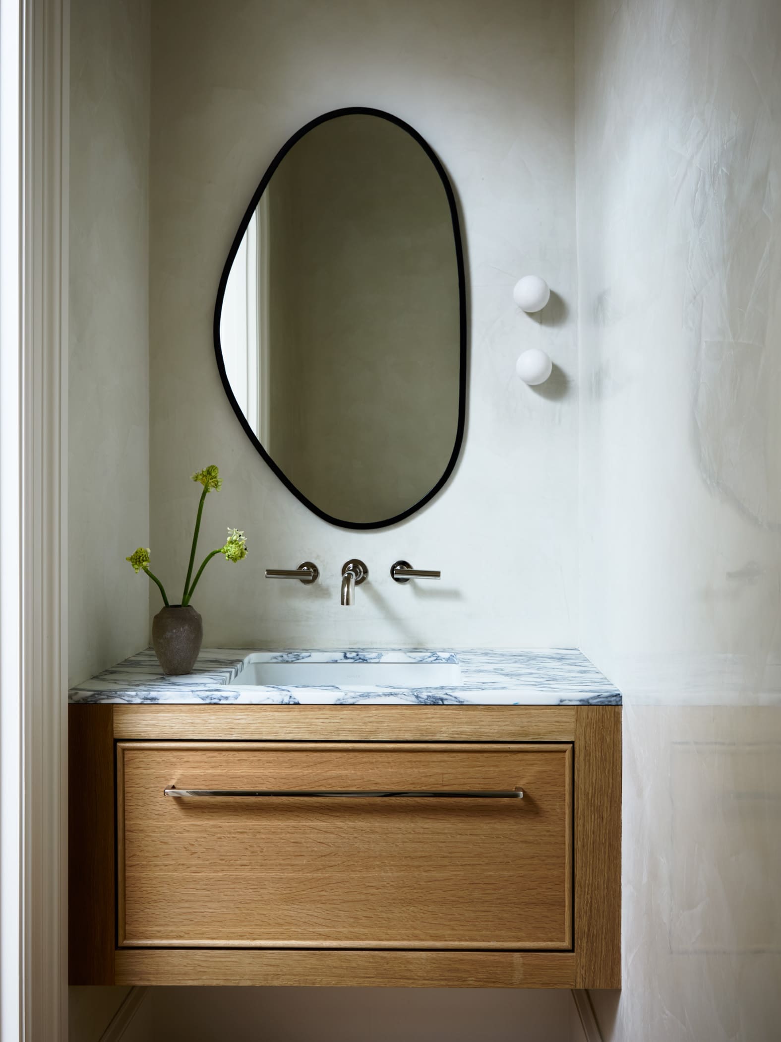 Round black-framed mirror above a marble countertop and wooden vanity, with a small vase of green stems on the left and soft bathroom lighting.