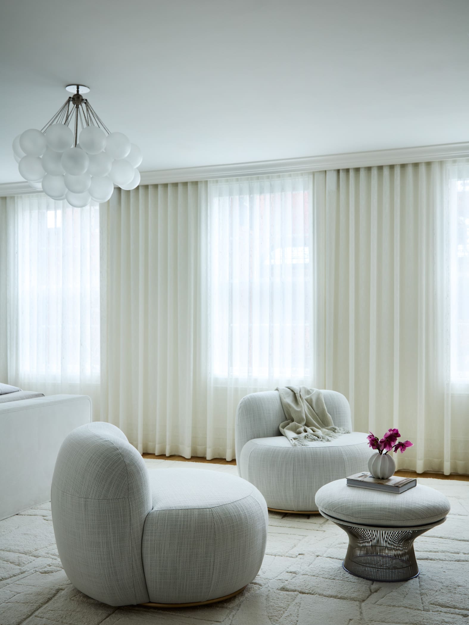 Living room with pale gray rounded ottomans, a glass-tiber coffee table, and sheer white curtains under a bubble chandelier.
