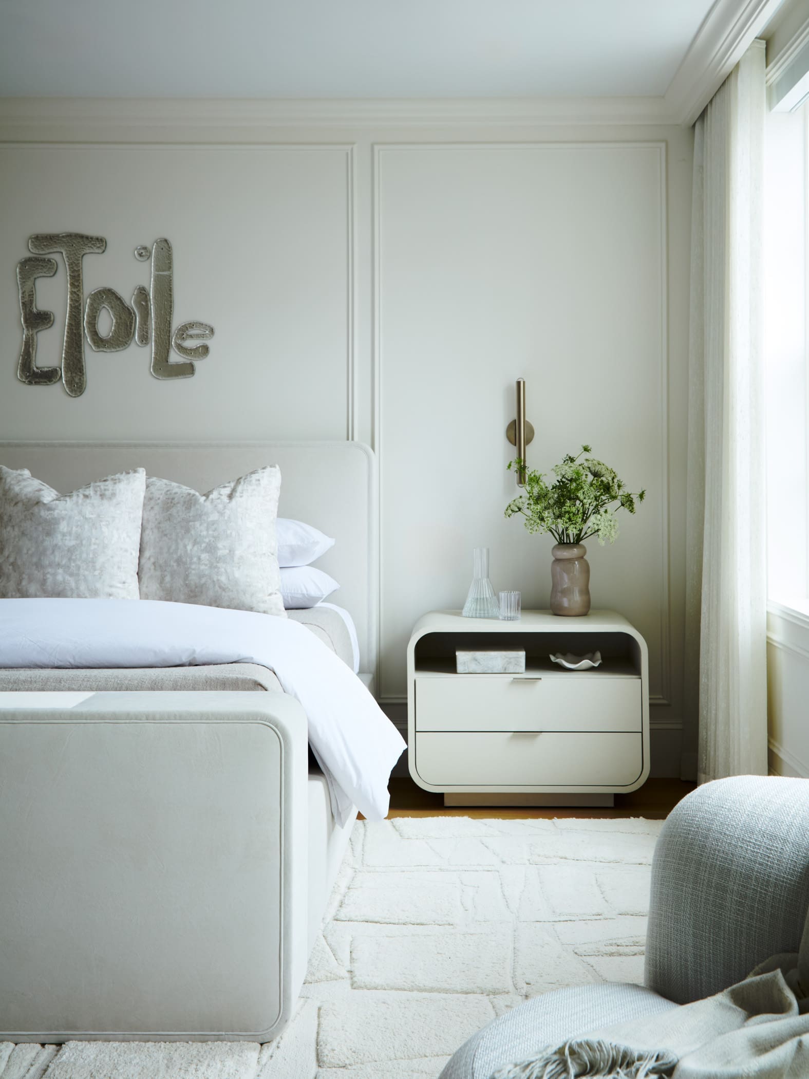 Neutral-toned bedroom with a pale bed, decorative pillows, and metallic word 'Elle' on the wall above the headboard.