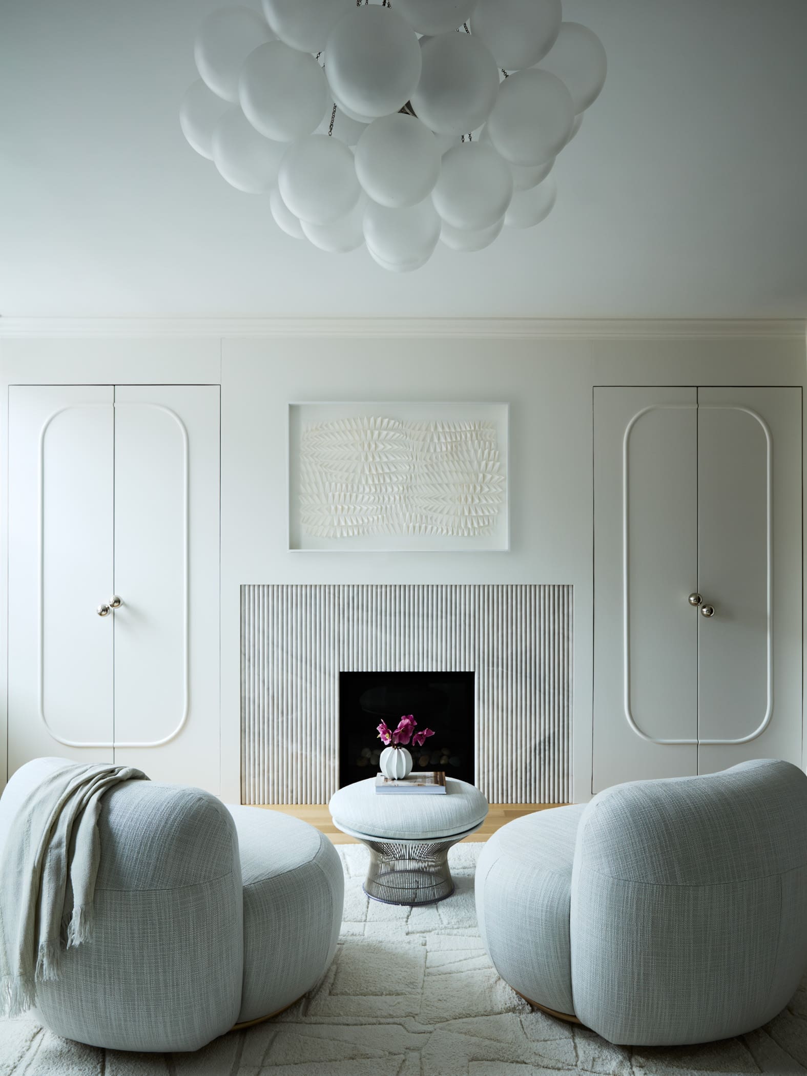 Light gray living room with round cushioned ottomans facing a built-in fireplace and pale walls, a small table with a pink orchid sits in the fireplace nook, and white bubble chandelier hangs overhead.
