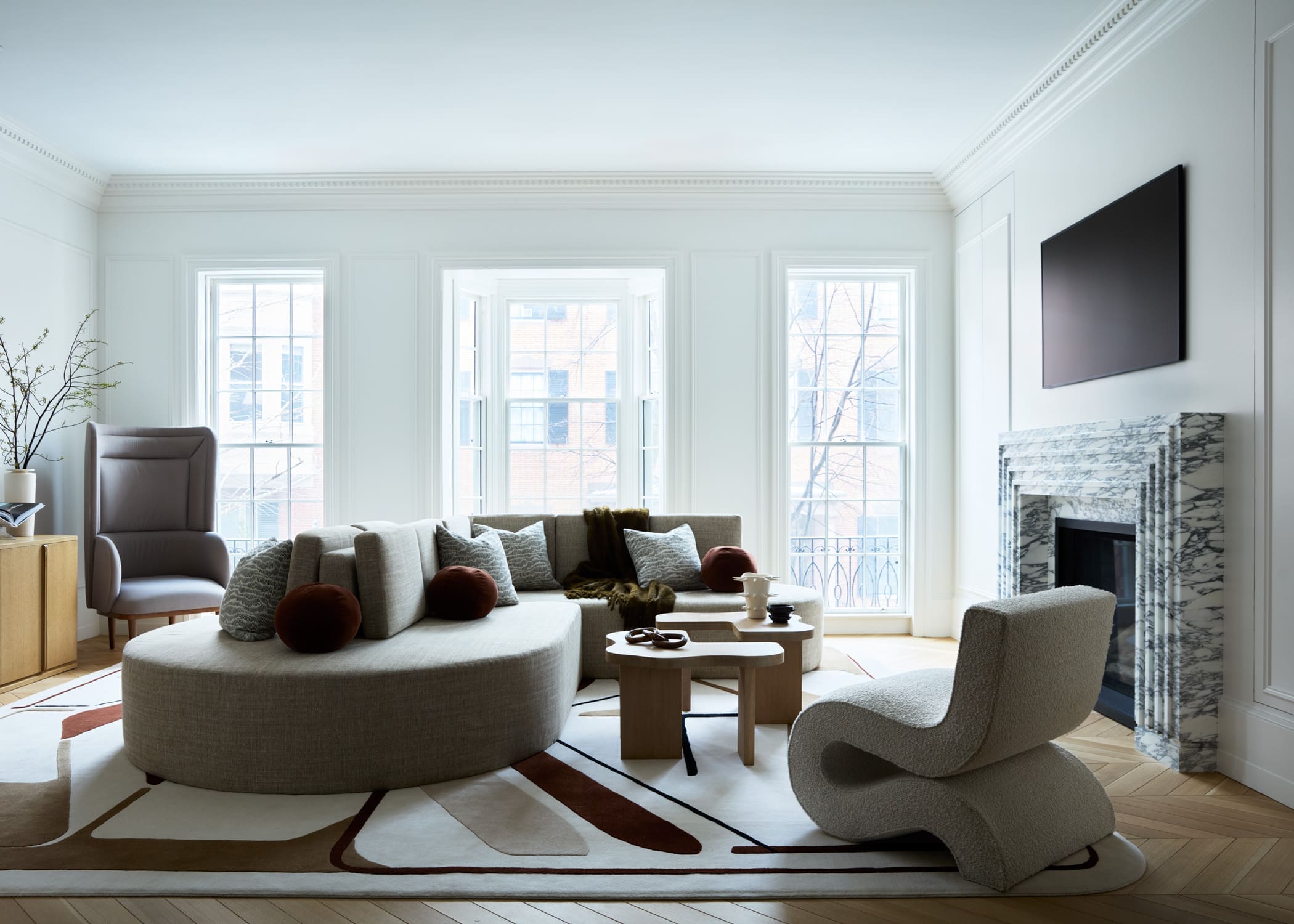 Bright living room with a curved beige sectional, round ottoman, and marble fireplace facing large windows, in soft daylight.