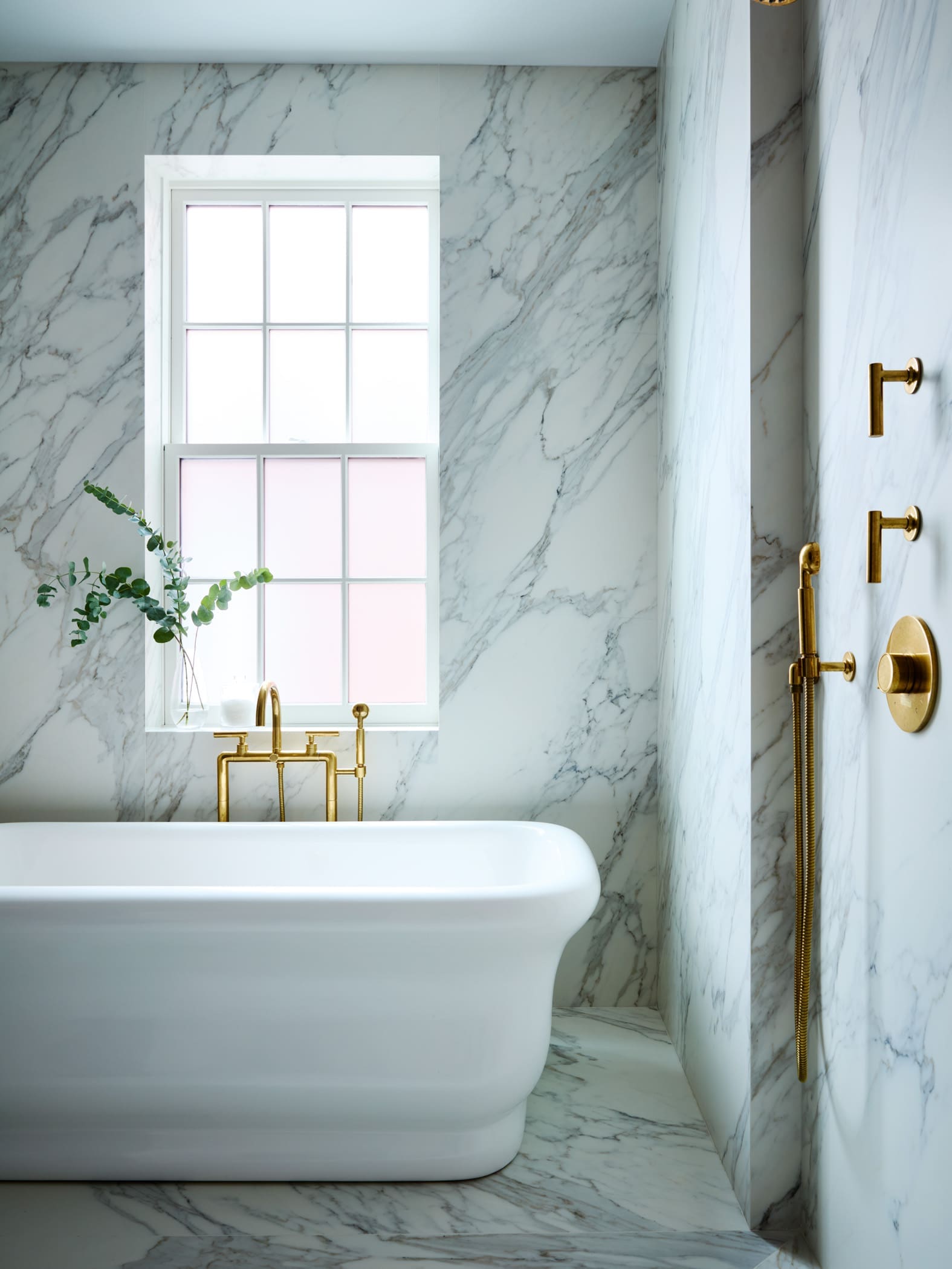 White freestanding bathtub in a marble bathroom with gold-tone fixtures and a large window letting in soft light.