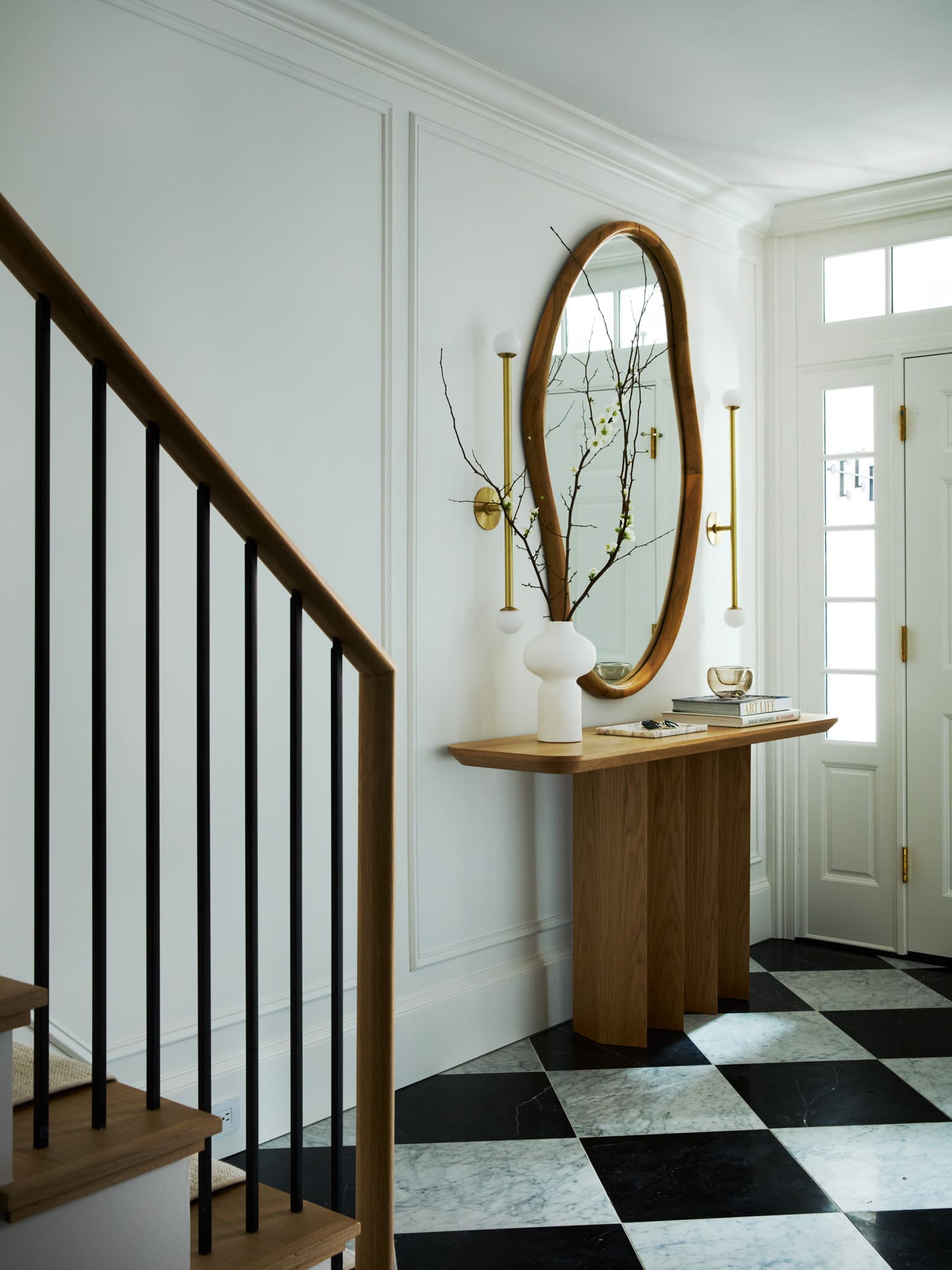 Entry hall with a wooden-framed oval mirror above a console table, a white vase with branches, and brass wall sconces by the front door.