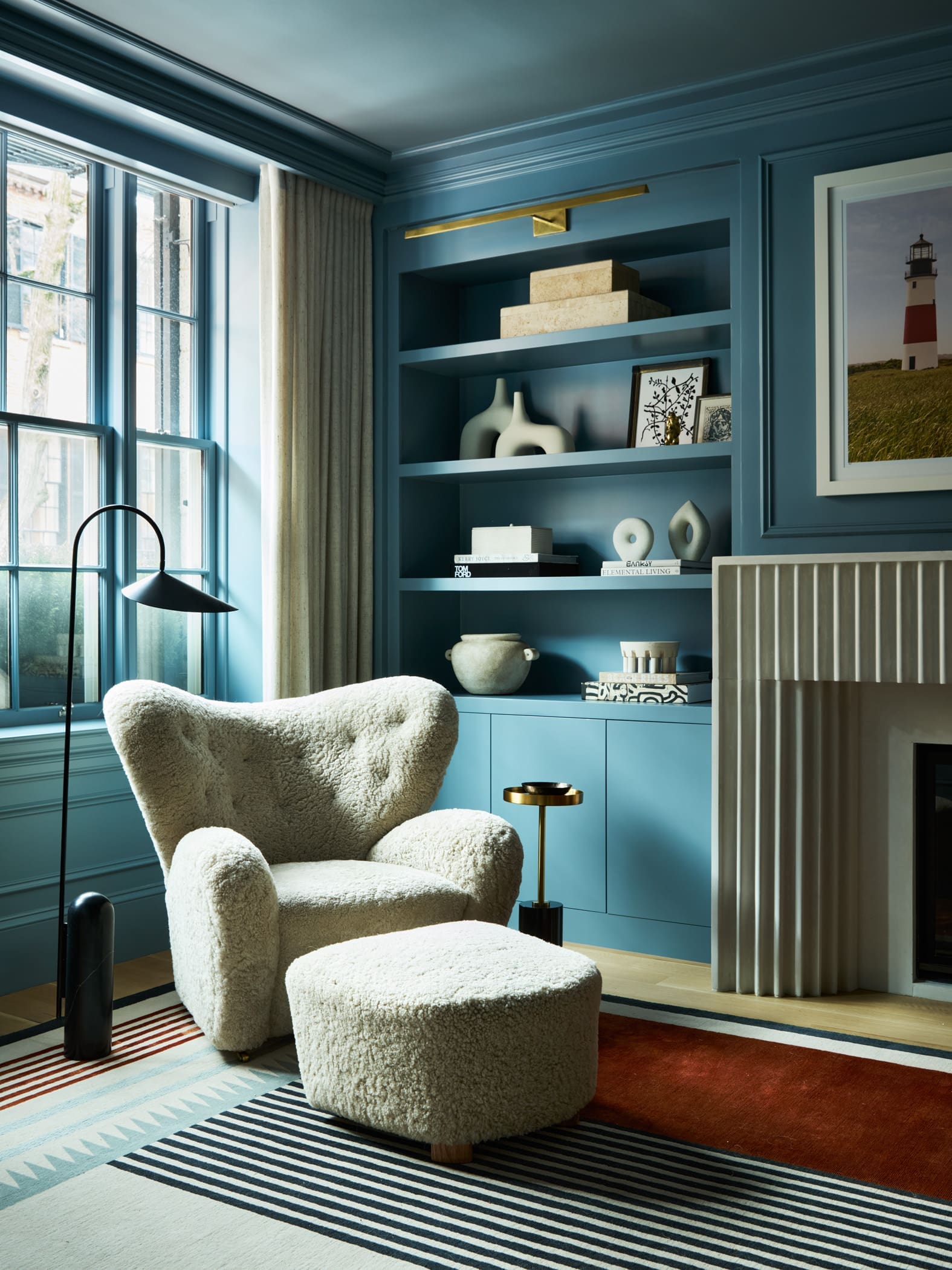 Cozy blue living room with a cream boucle chair and ottoman, built-in shelves, and a striped rug near a large window.