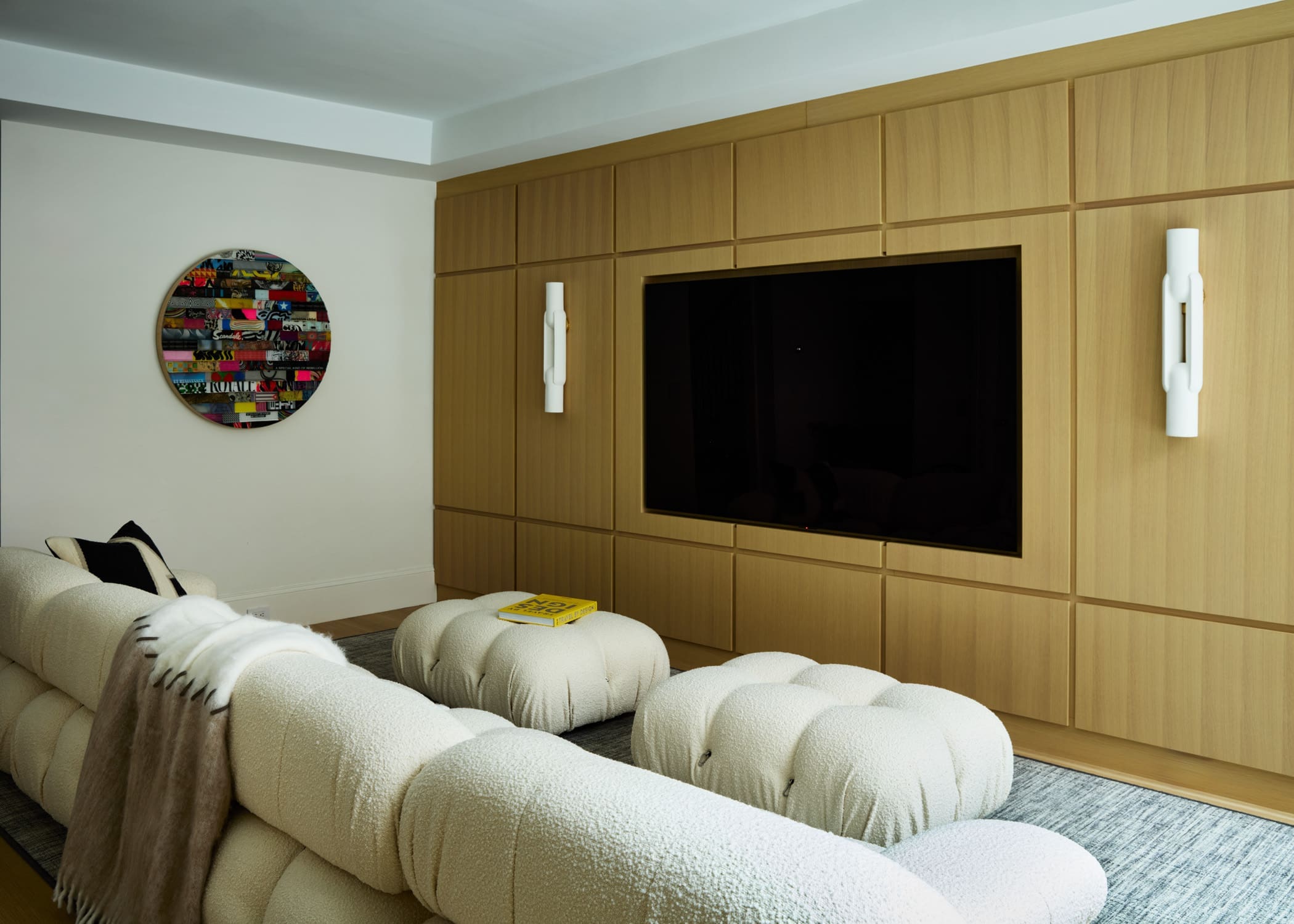 Living room with a wood-paneled wall unit housing a flat-screen TV, white tufted seating, and a colorful round collage wall art on the pale wall.