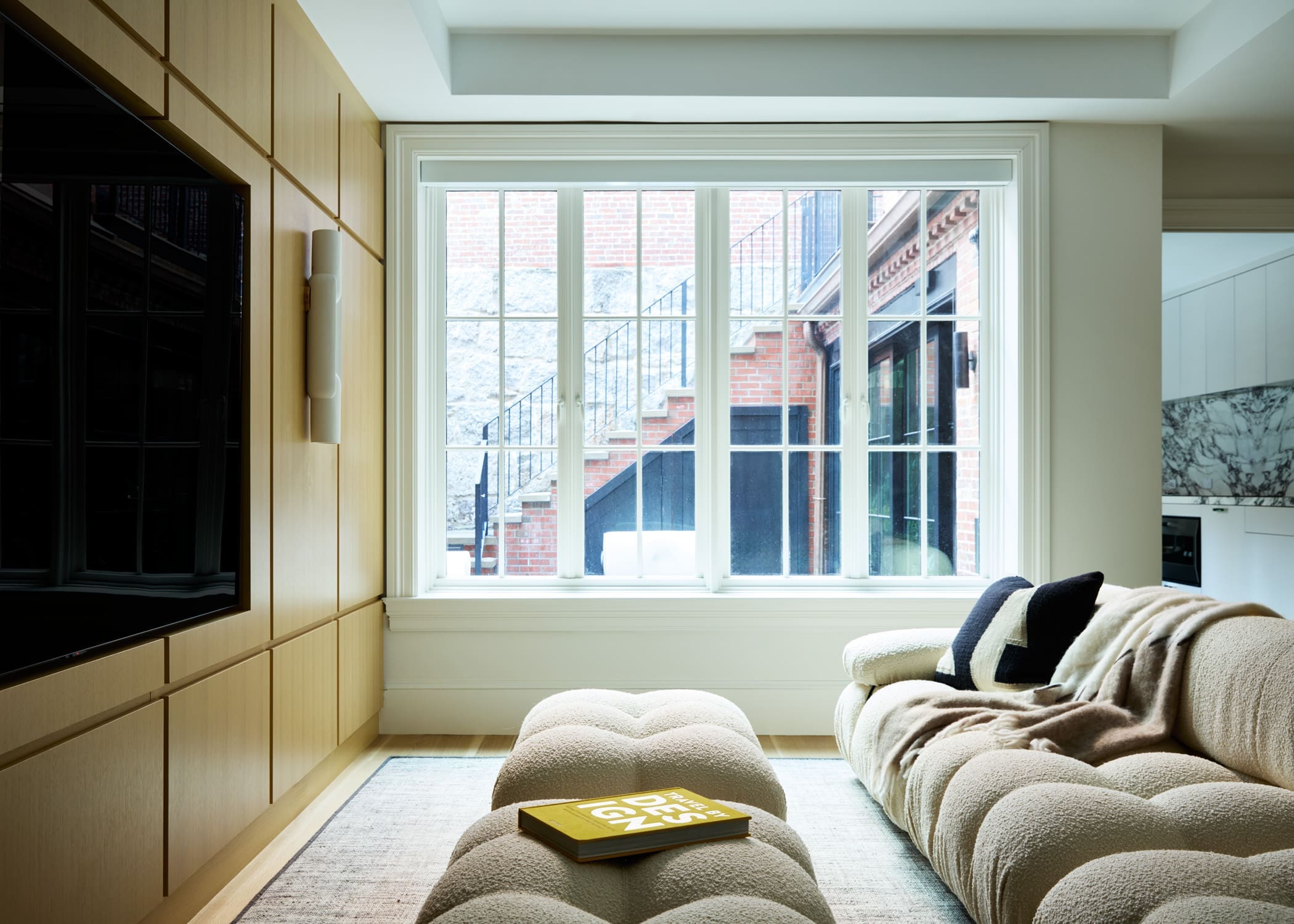 Living room with floor-to-ceiling wooden cabinets on the left, a large window showing brick exterior and stairs, and a beige tufted sofa with a book on a small ottoman in the foreground.