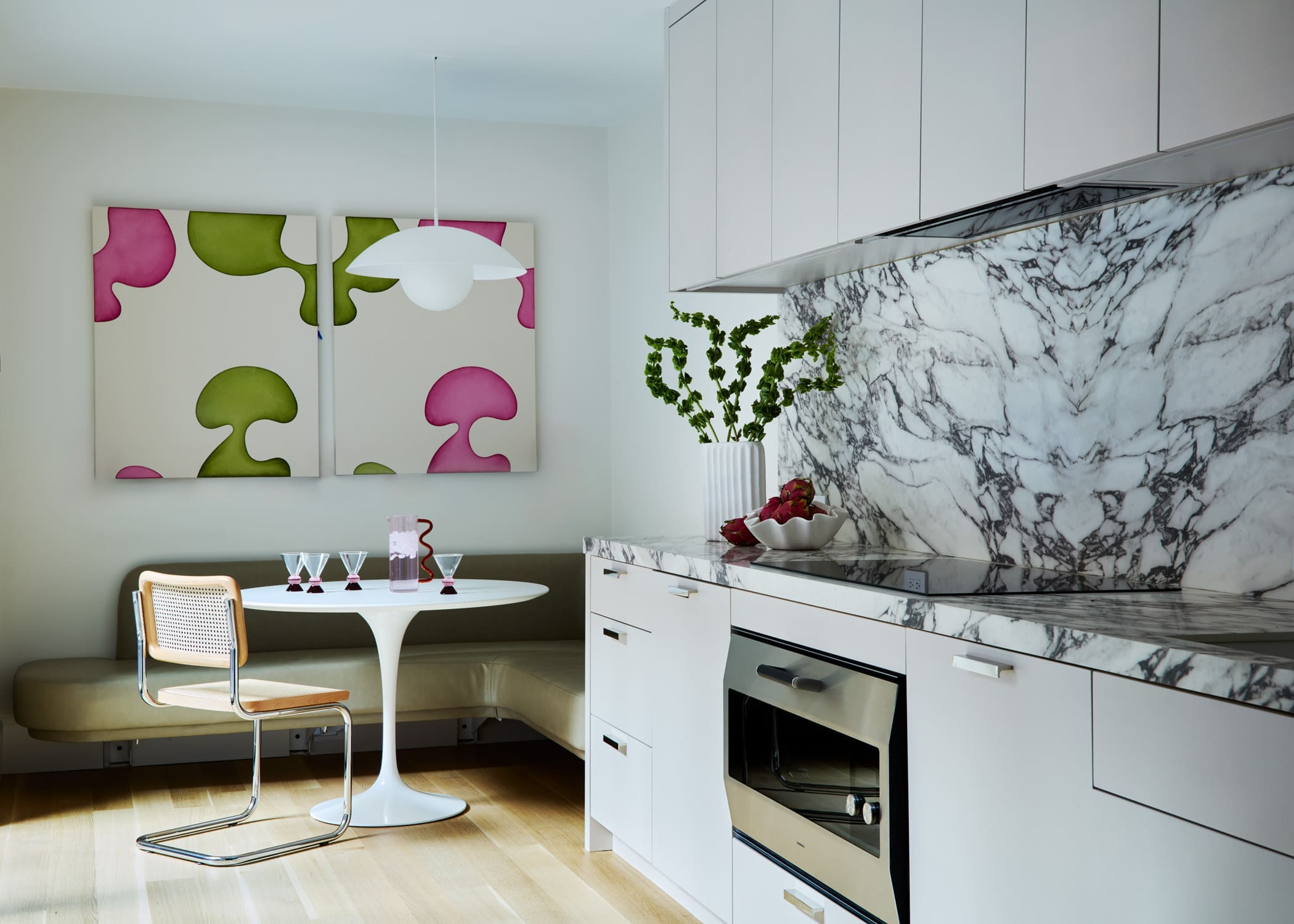 Modern kitchen with white cabinets, marble backsplash, and a round white table set with pink drinks and champagne glasses; abstract pink‑green wall art in the background.