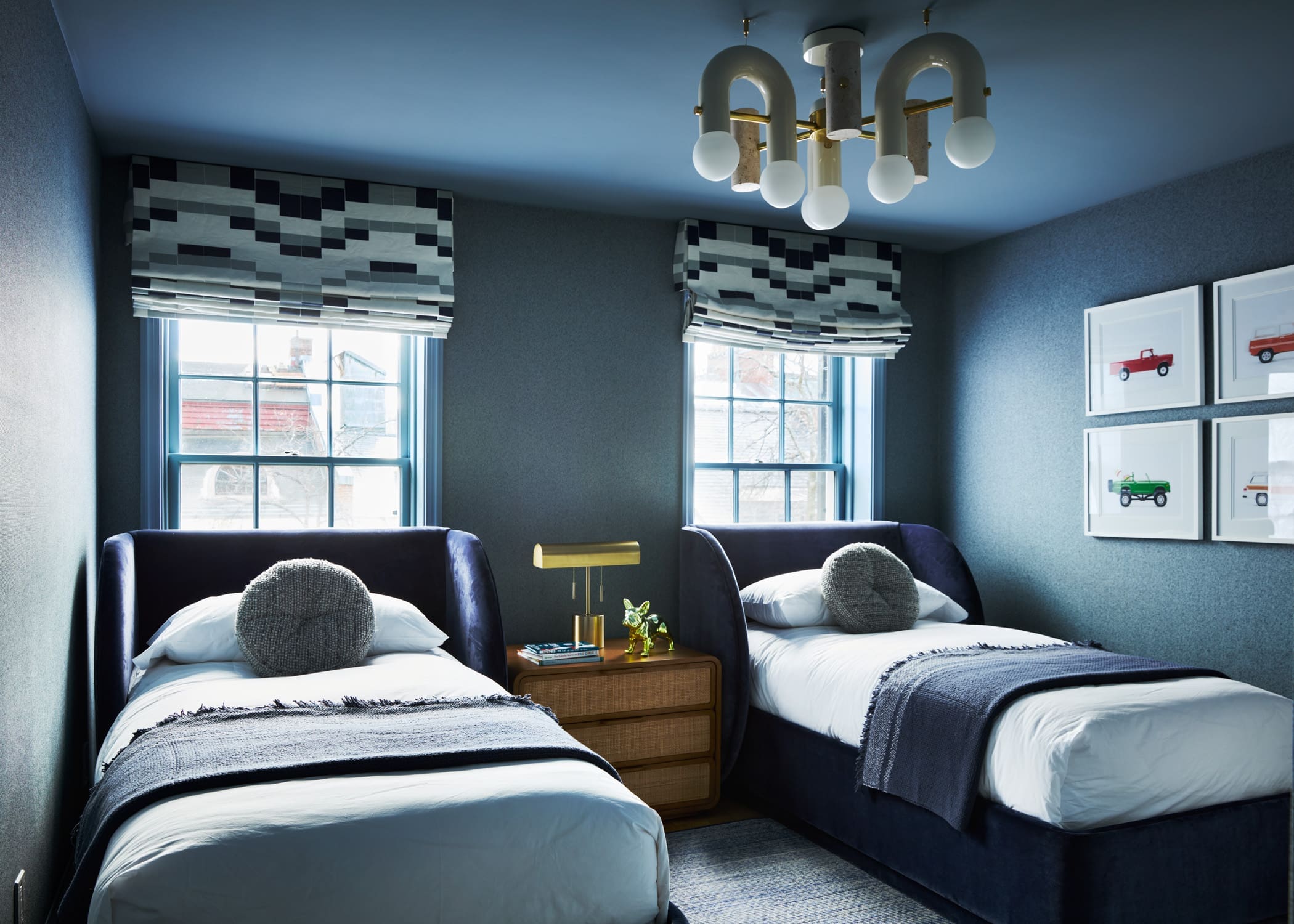 Twin beds in a blue-toned bedroom with a wooden nightstand, brass lamp, and geometric window shades