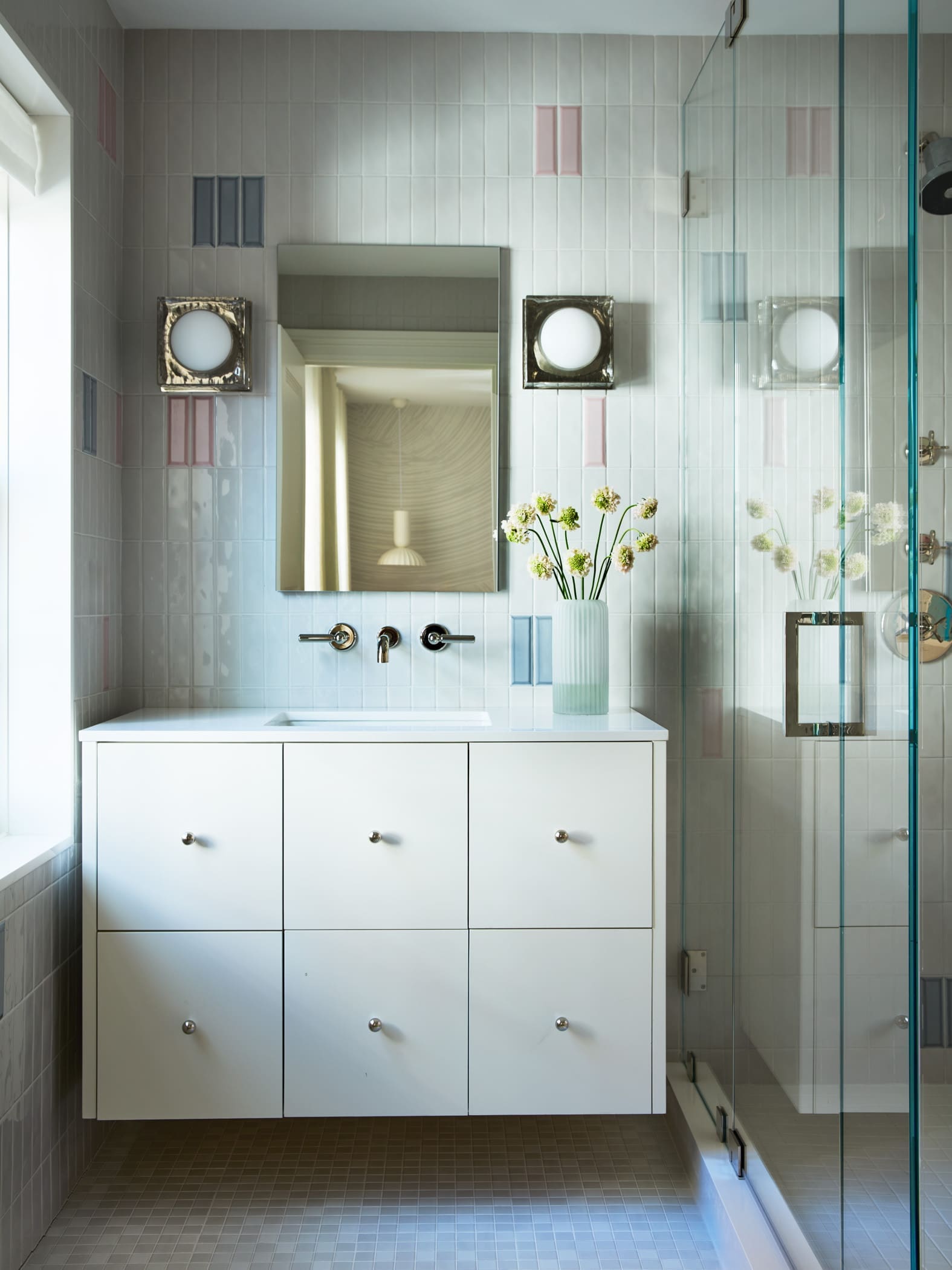 White bathroom vanity with six drawers and a rectangular mirror, flanked by two square wall lights, next to a glass shower enclosure.
