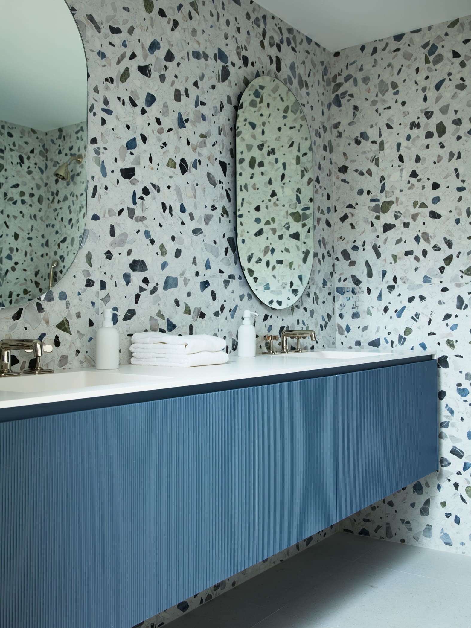 Modern bathroom vanity with blue ribbed cabinets, terrazzo walls, and oval mirrors above a white countertop with two sinks and soap dispensers.