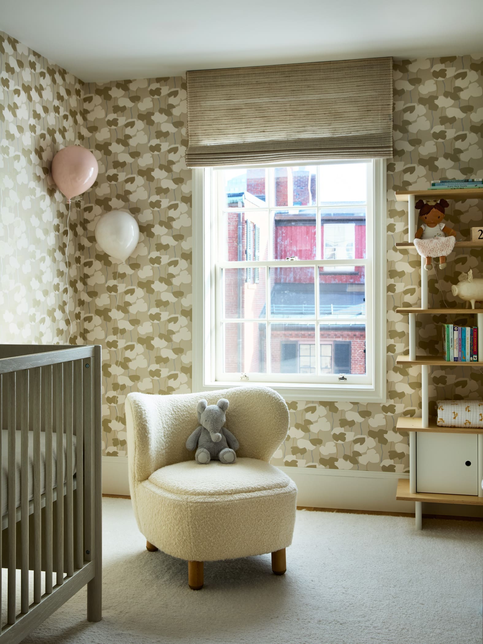 Cozy nursery corner with a cream upholstered chair and stuffed bear, floral wallpaper, a window with a striped shade, and a shelf of toys and books.