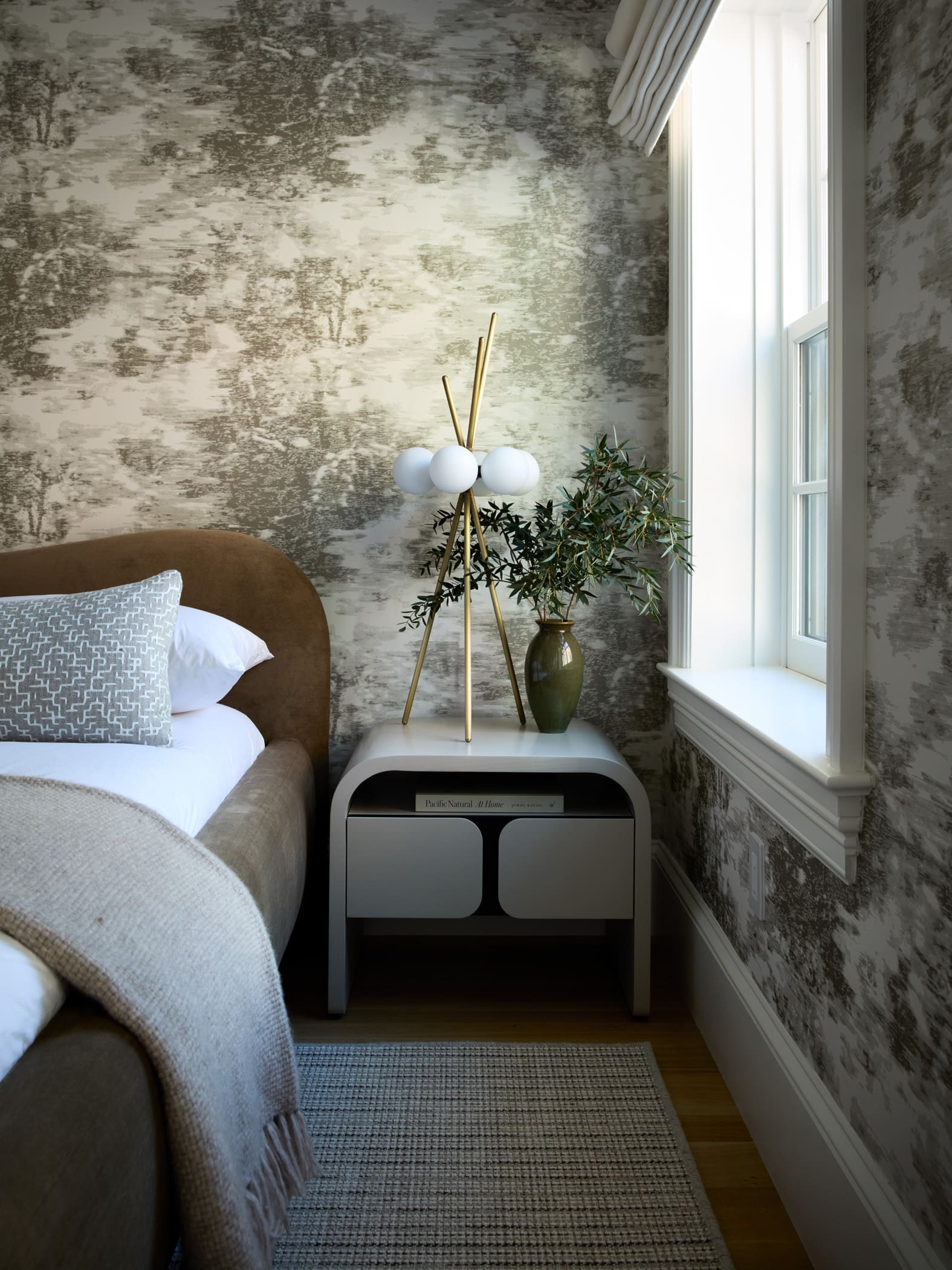 Cozy bedroom corner with a brown upholstered bed, gray throw blanket, and a patterned pillow, next to a modern gold tripod lamp and a green plant by a sunny window.