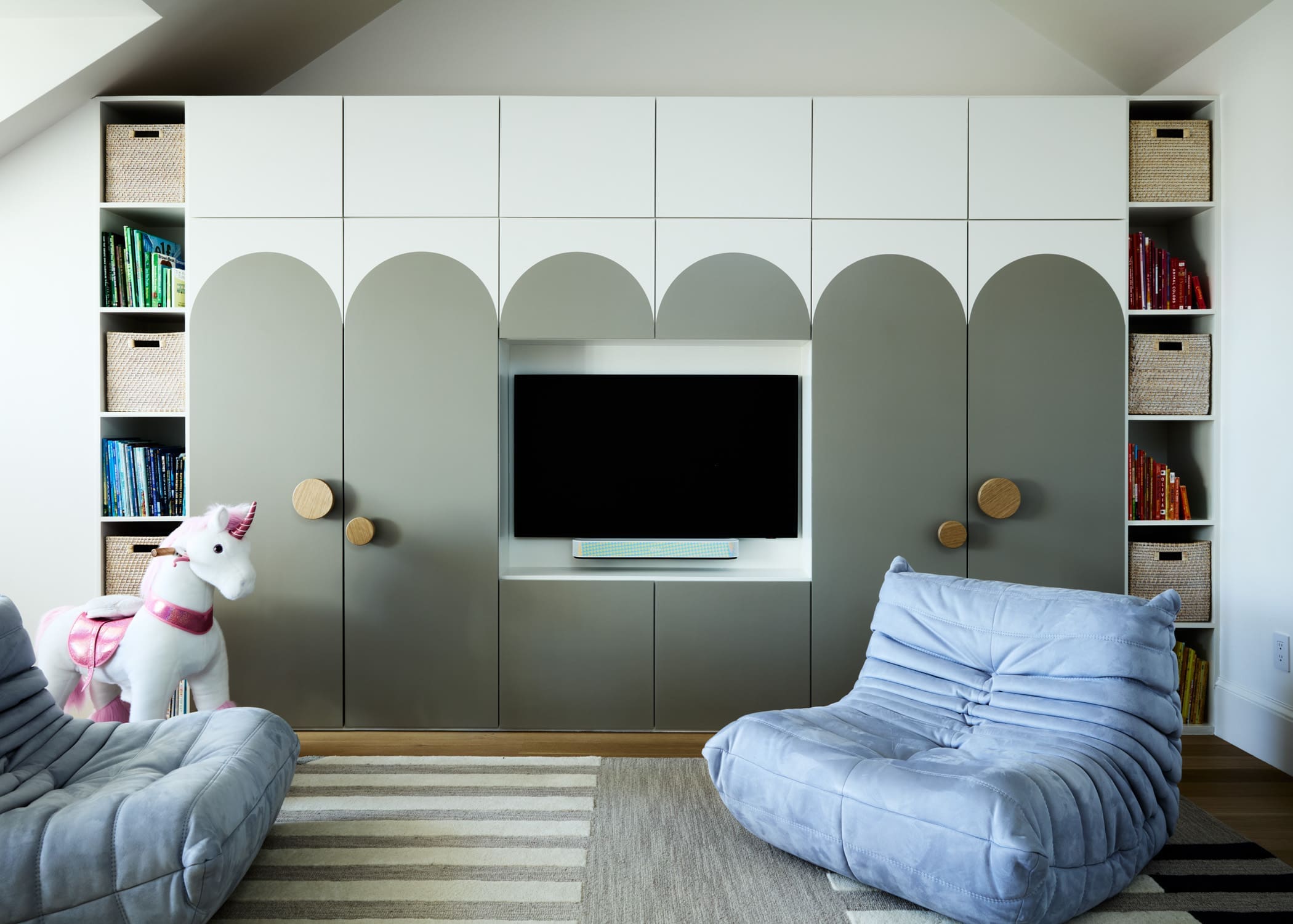 Modern living room storage wall with grey cabinets, woven baskets, and shelves holding books; TV centered with a small decorative shelf below. A pink-and-white unicorn plush sits on a blue beanbag chair in the foreground.