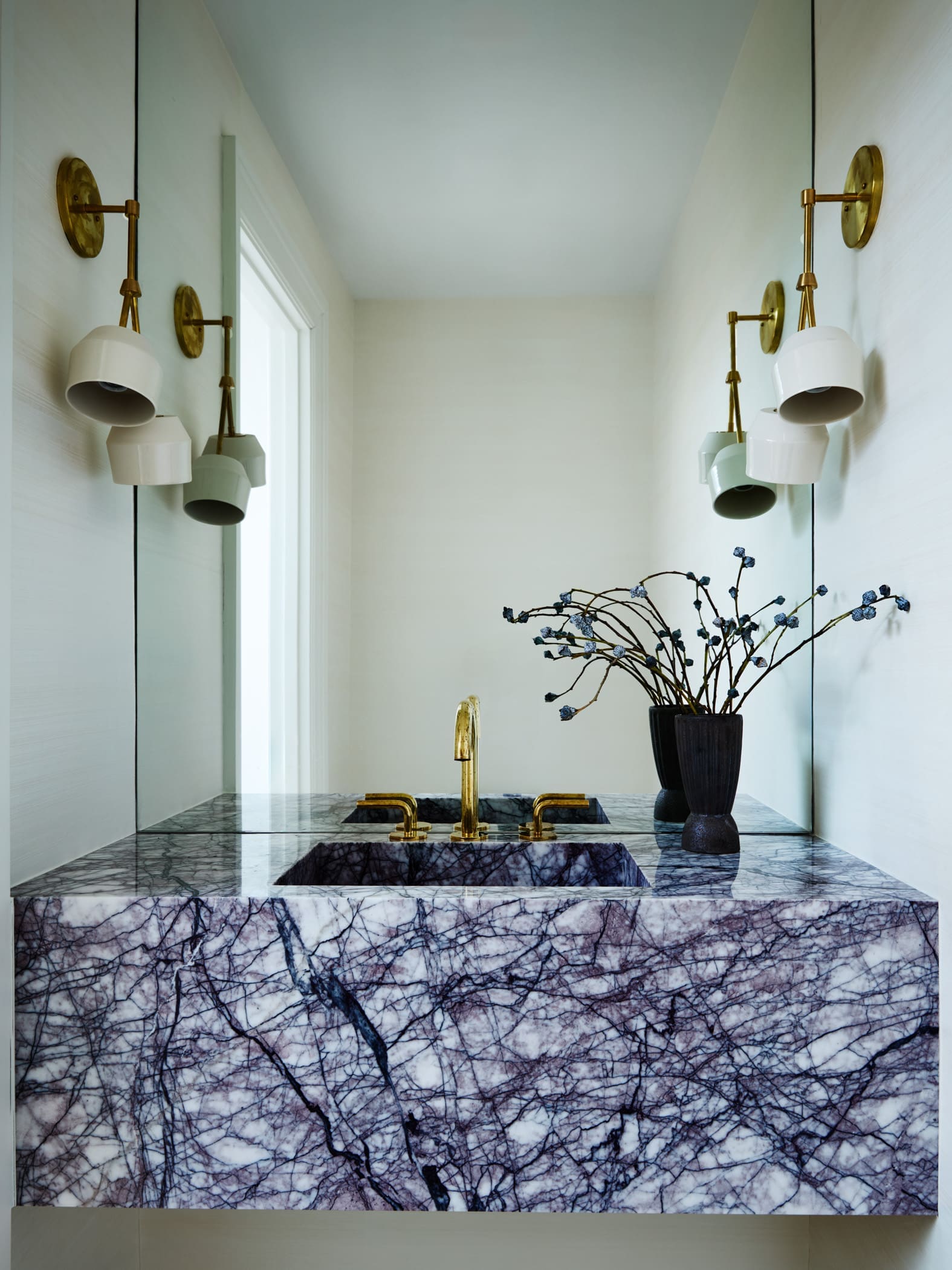 Symmetrical bathroom vanity featuring a white marble countertop with dark veining, gold faucet, and a vase of decorative branches on the right, framed by brass wall sconces and mirrors.
