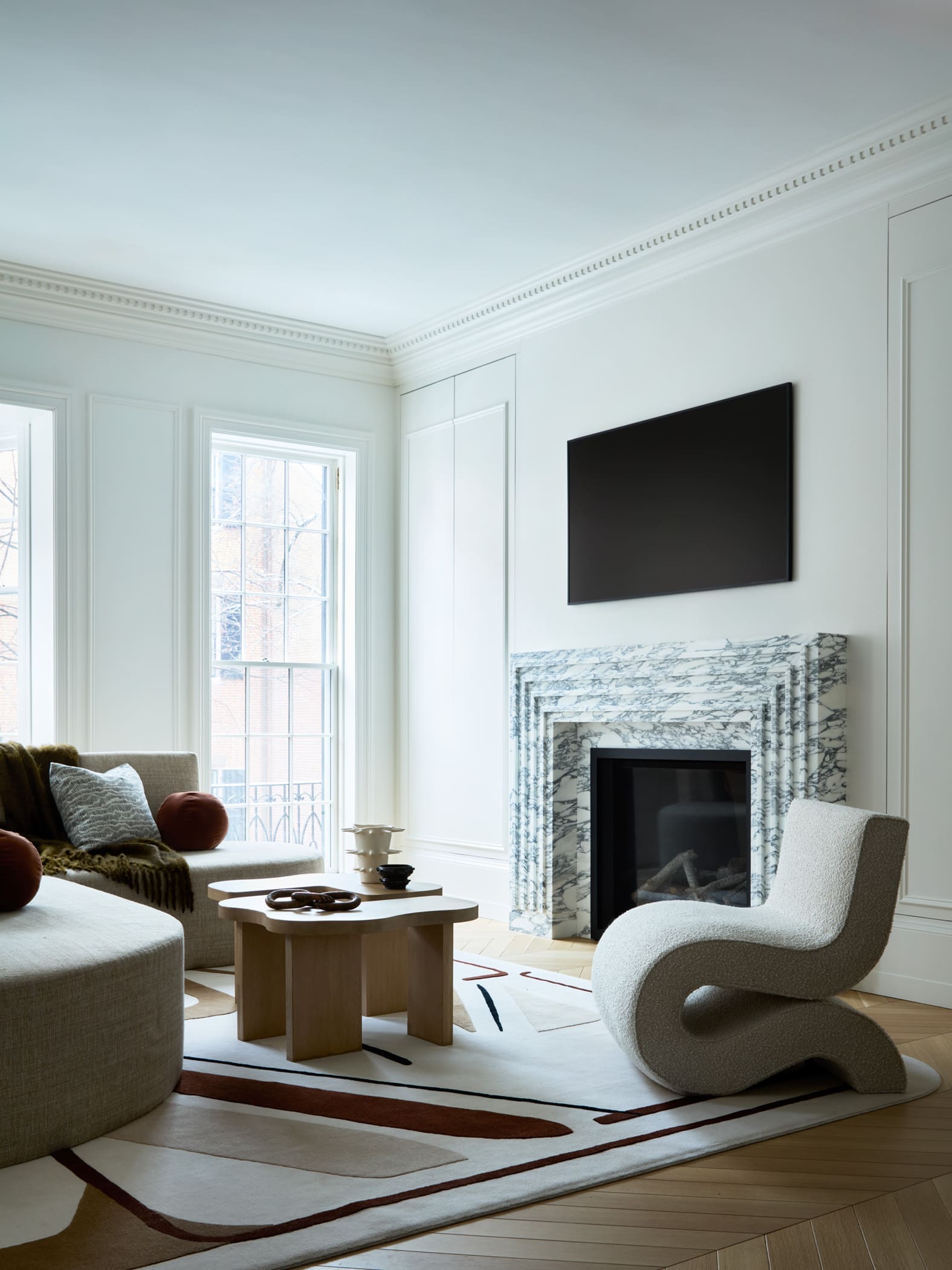 Bright living room with white paneled walls, large windows, and a marble fireplace topped by a wall-mounted TV.