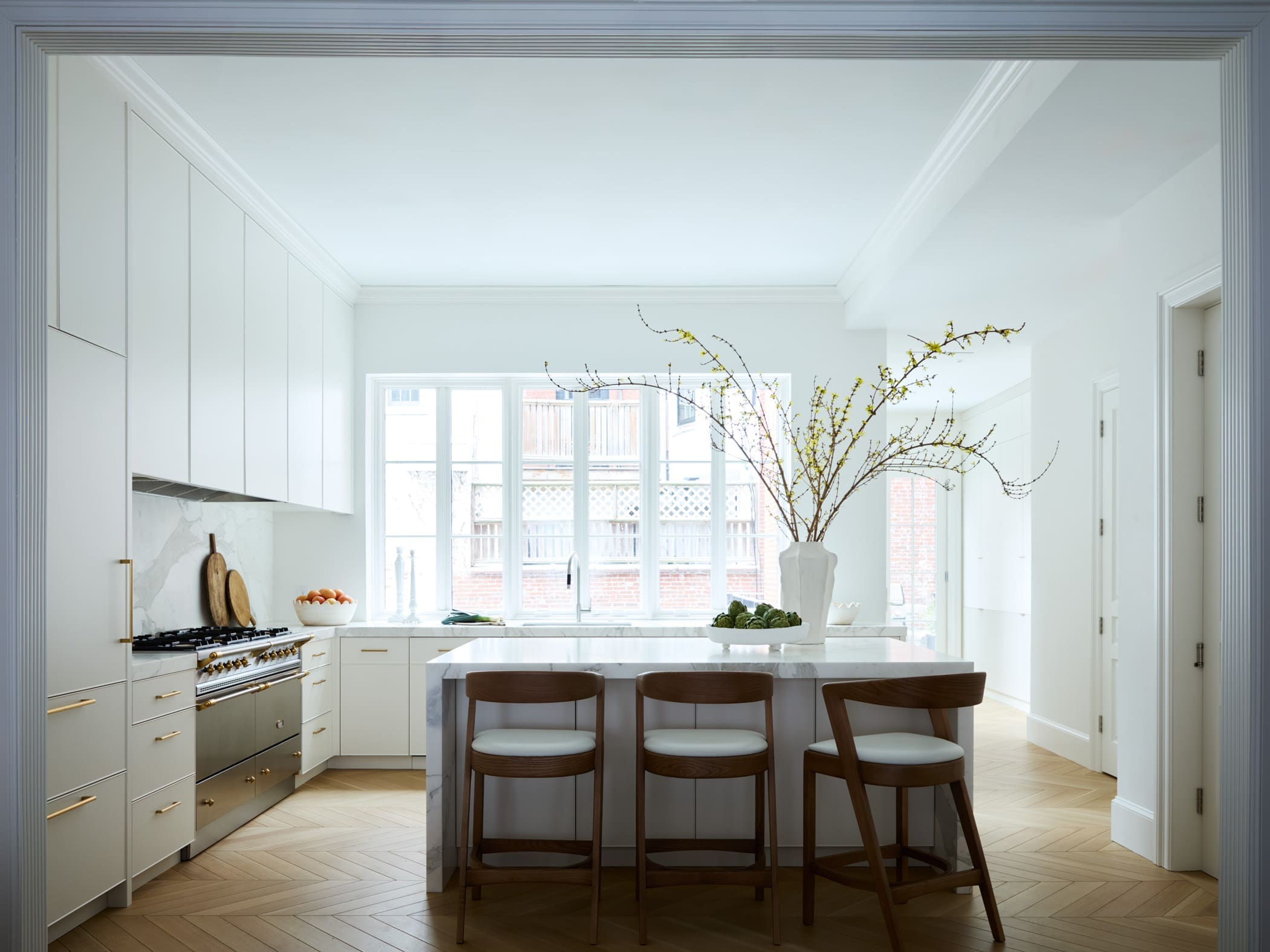 Bright white kitchen with marble island, wooden bar stools, and a vase of branches by a large window.