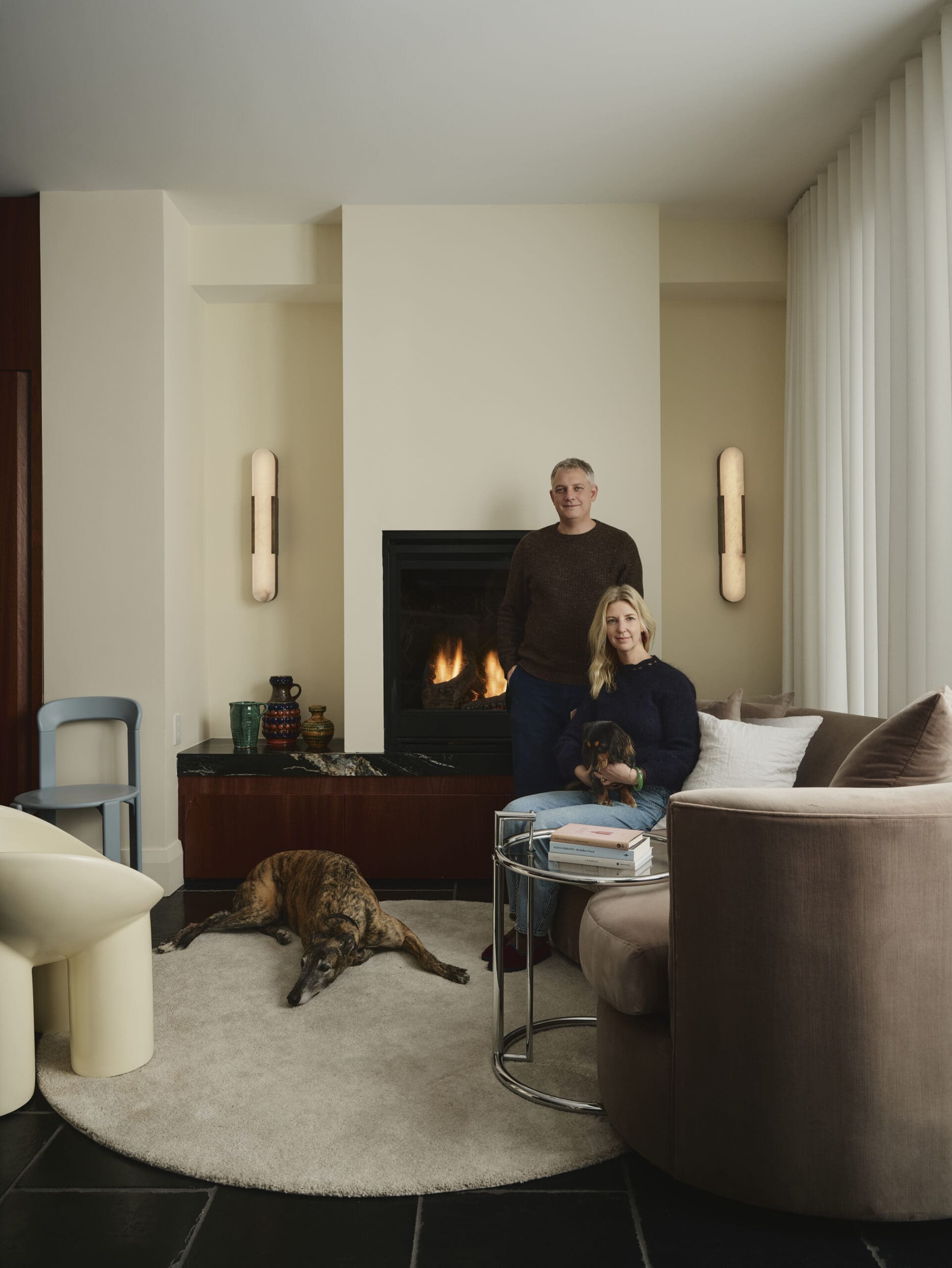 Man standing behind a seated woman with two dogs in a cozy living room featuring a lit fireplace and large windows with vertical blinds.