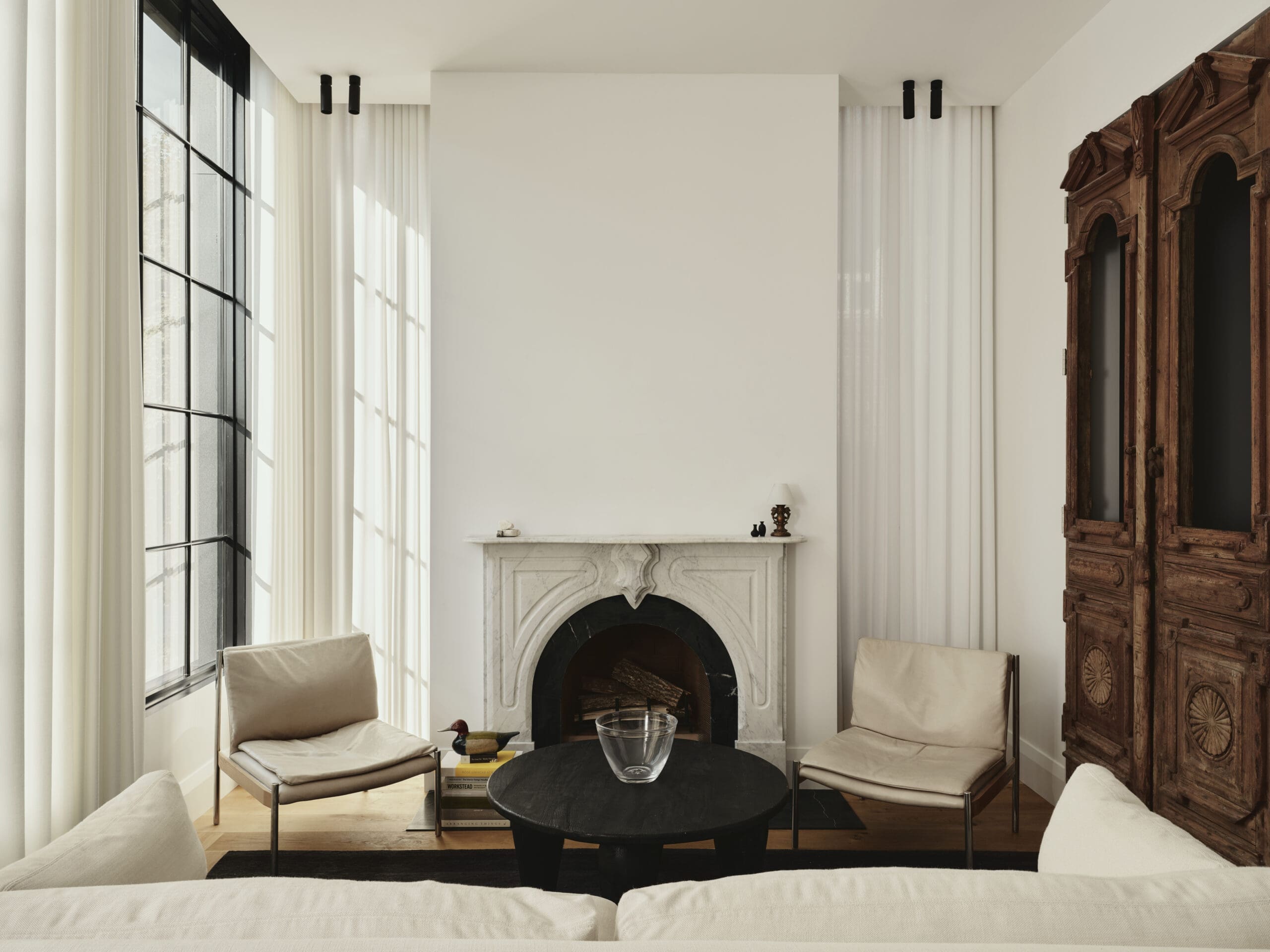 Bright living room with tall windows and white curtains, a marble fireplace, a black round coffee table, and cream chairs.