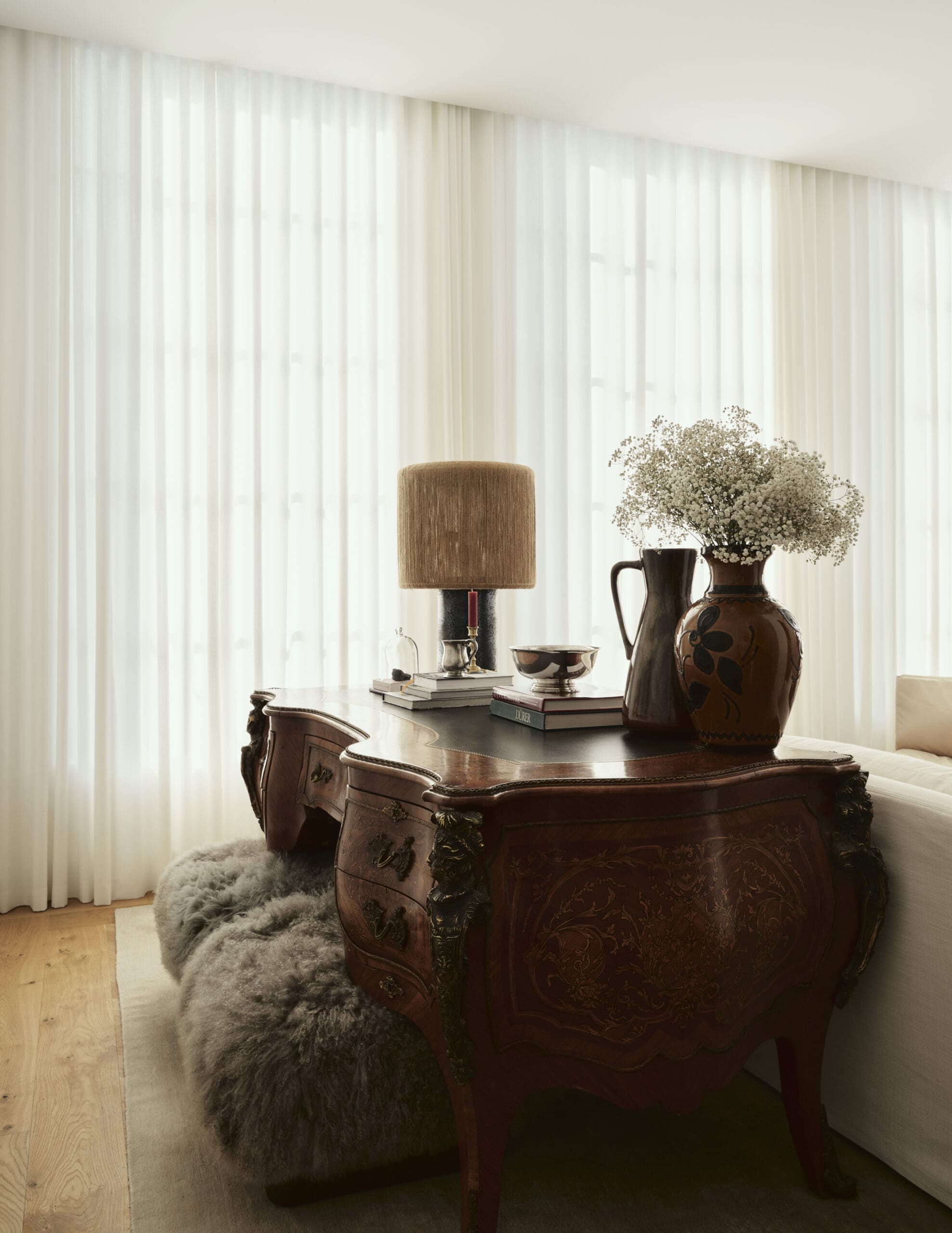 Antique carved wooden desk with a tall beige lampshade, a vase of white baby's-breath flowers, and stacked books against sheer white curtains.
