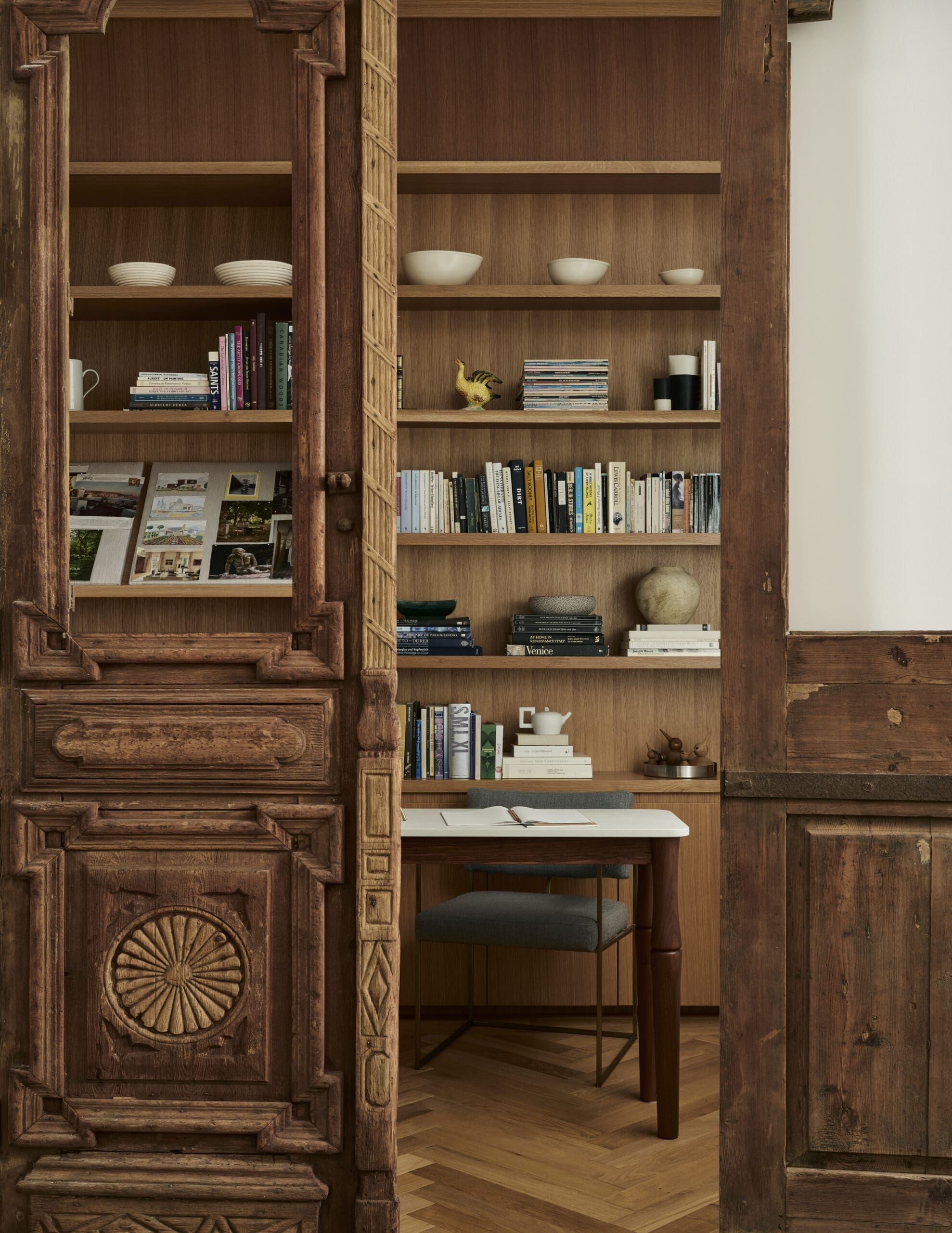 Carved wooden cabinet with open shelves holding books, photos, bowls, and a small desk with a notebook and chair underneath.